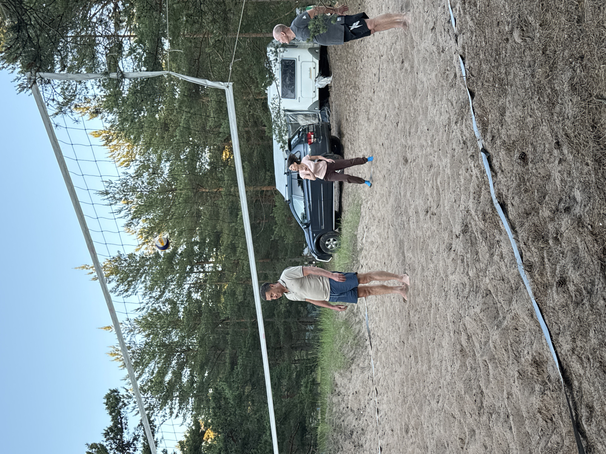 A group of three adults are playing volleyball on a sandy court surrounded by pine trees. A volleyball is in the air above the net. A dark-colored SUV and a white camper trailer are parked behind the court. The setting appears to be a recreational area in Ozerki (Krasnodolinskaya), Russia, likely a campsite or park. The lighting suggests it is late afternoon or early evening. The man closest to the camera is wearing a light-colored polo shirt and blue shorts. A woman in the middle is wearing a pink shirt and brown pants, and appears to be mid-action. The man on the far right is wearing a grey t-shirt and black shorts, and is holding a small bunch of greenery. The sand on the court is uneven, and a thin blue and black tape marks the boundary.