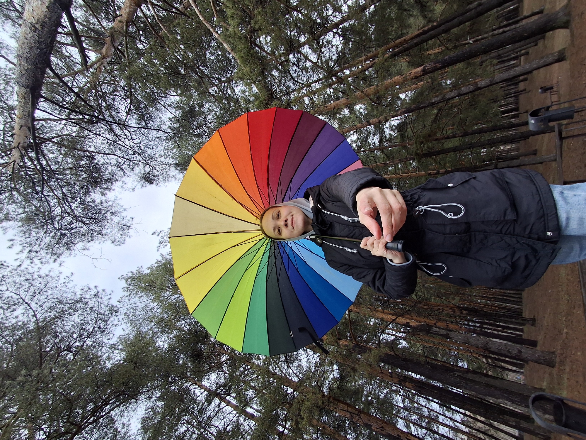 A young person stands outdoors in a forest, holding a large, vibrant rainbow-colored umbrella above their head. The person, who appears to be female, is looking directly at the camera with a slight smile and has their right arm extended forward, pointing an index finger playfully towards the viewer. They are dressed in a dark winter jacket over a light grey hooded top.
The background is dominated by a dense forest, featuring numerous tall, slender tree trunks with dark bark and a canopy of pine-like foliage. The ground beneath the trees is covered with brown earth and fallen needles. The sky above is overcast and a light grey, indicating a cloudy daytime. In the mid-ground on the right, some dark metal structures, possibly outdoor furniture or equipment, are partially visible among the trees.
The image conveys a cheerful and casual atmosphere, with the bright, multi-colored umbrella providing a striking contrast to the muted, natural tones of the forest on an overcast day. This scene is set in a forest in Chaikovsky, Russia.