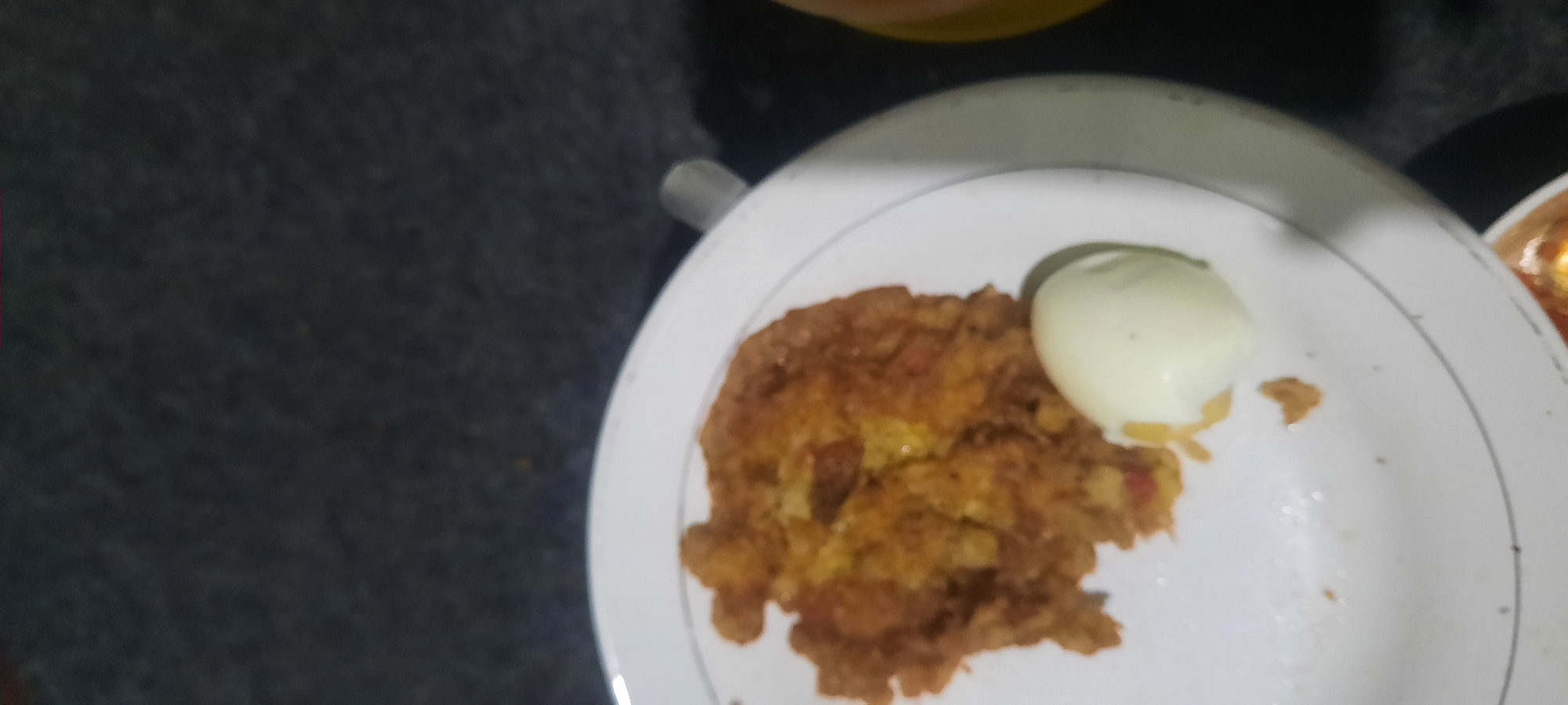 A close-up, slightly blurry shot shows a plate of food on a dark, textured surface. The plate is white with a thin, silver-colored rim. On the left side of the plate is a piece of browned food, possibly an omelet or fritter, with visible red and yellow pieces mixed in. To the right of this is a whole, peeled hard-boiled egg. The background is out of focus, appearing as a dark, fuzzy texture. The scene is likely indoors, with the lighting suggesting an artificial light source. There is no indication of a specific location in North Jakarta, Indonesia.