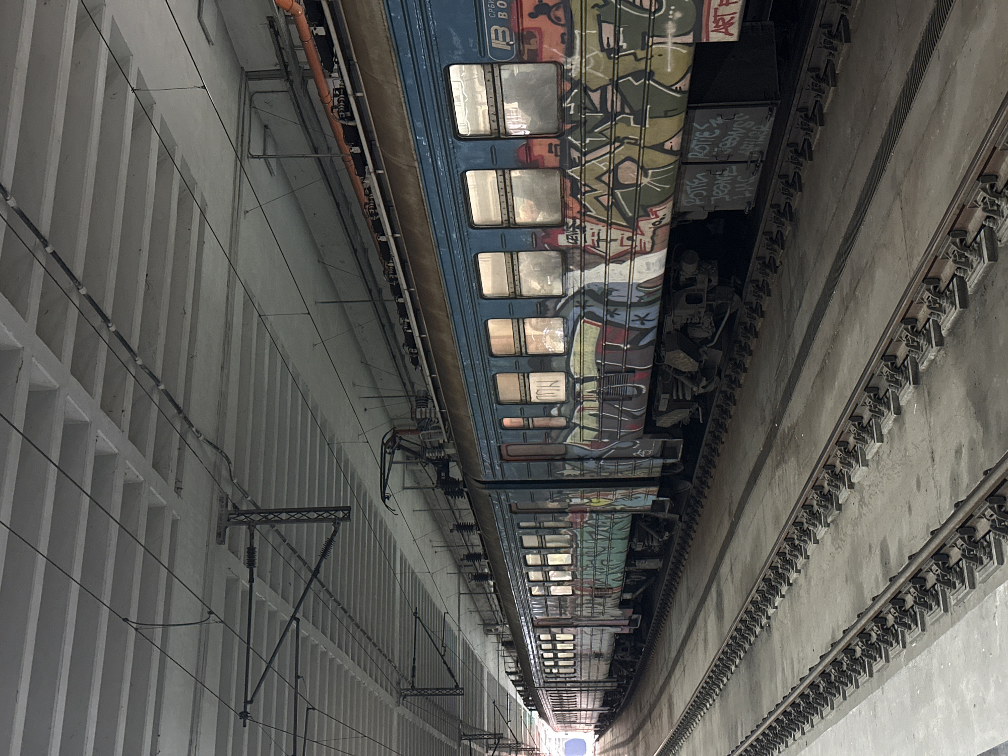 The image shows a train car in a tunnel in Belgrade, Serbia. The train is covered in graffiti. The tunnel has white walls on one side and gray walls on the other. The tunnel appears to be a subway.