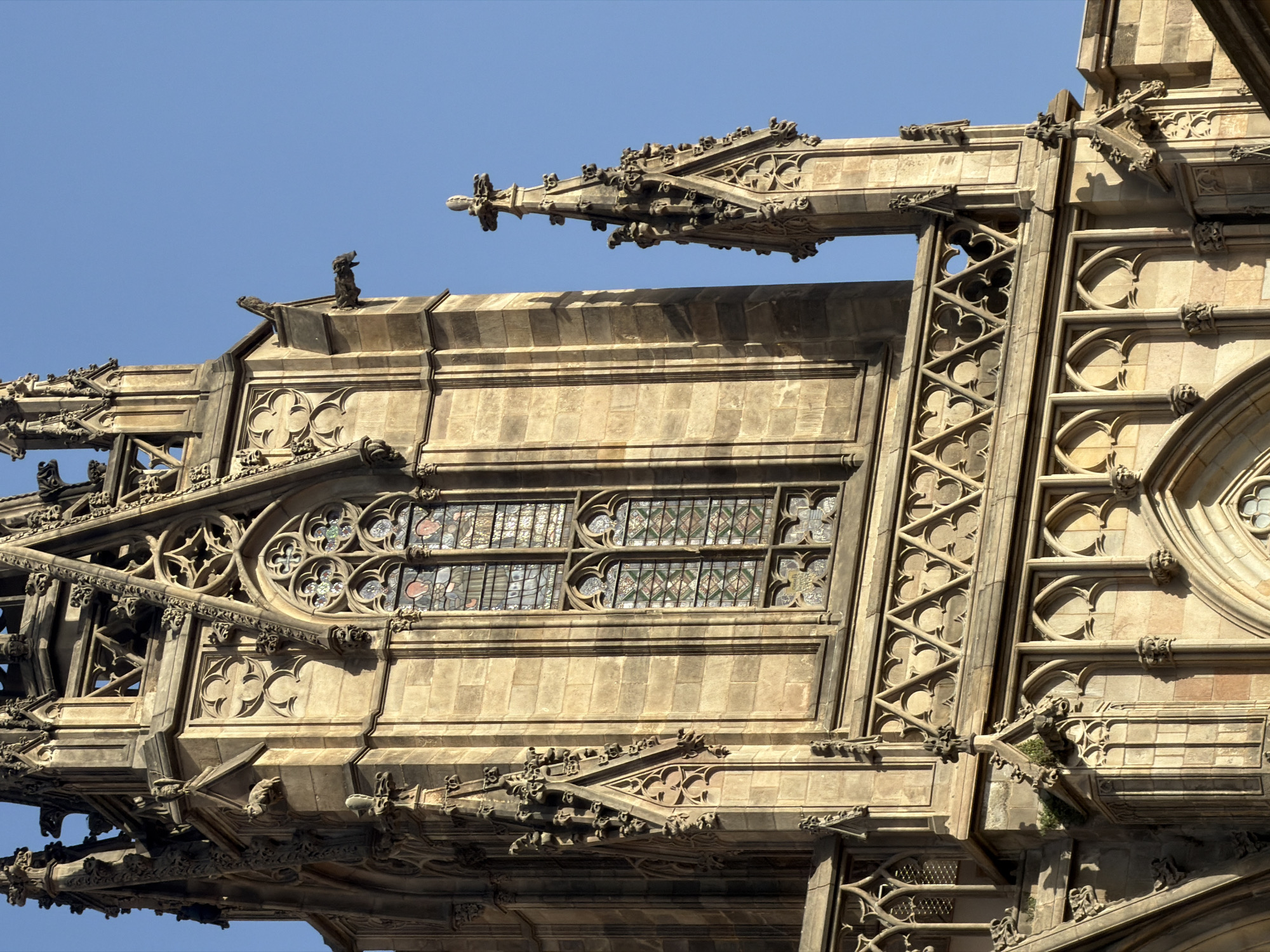 The photograph captures a close-up view of a segment of the Barcelona Cathedral's facade, specifically its Gothic architecture, against a clear blue sky. The image focuses on the intricate details of the stone carvings, stained-glass windows, and decorative elements.
The primary subject is the upper portion of a tower or spire, characterized by its textured, weathered stone construction. Several gargoyles, designed as grotesque figures, are visible projecting from the building's edges, likely serving as decorative elements and drainage spouts. Two large, arched stained-glass windows are a prominent feature, showcasing geometric patterns and colored glass fragments. Below these windows, a decorative stone frieze runs horizontally, composed of repeated quatrefoil motifs enclosed within triangular frames. Further down, the facade displays a series of smaller, pointed archways and more carved ornamentation, including what appear to be small sculpted heads. A tall, slender pinnacle, topped with a cross-like finial, rises to the right of the main tower. The lighting suggests it is daytime, with strong sunlight casting shadows that highlight the three-dimensional aspects of the carvings. No people or active elements are present in the scene.