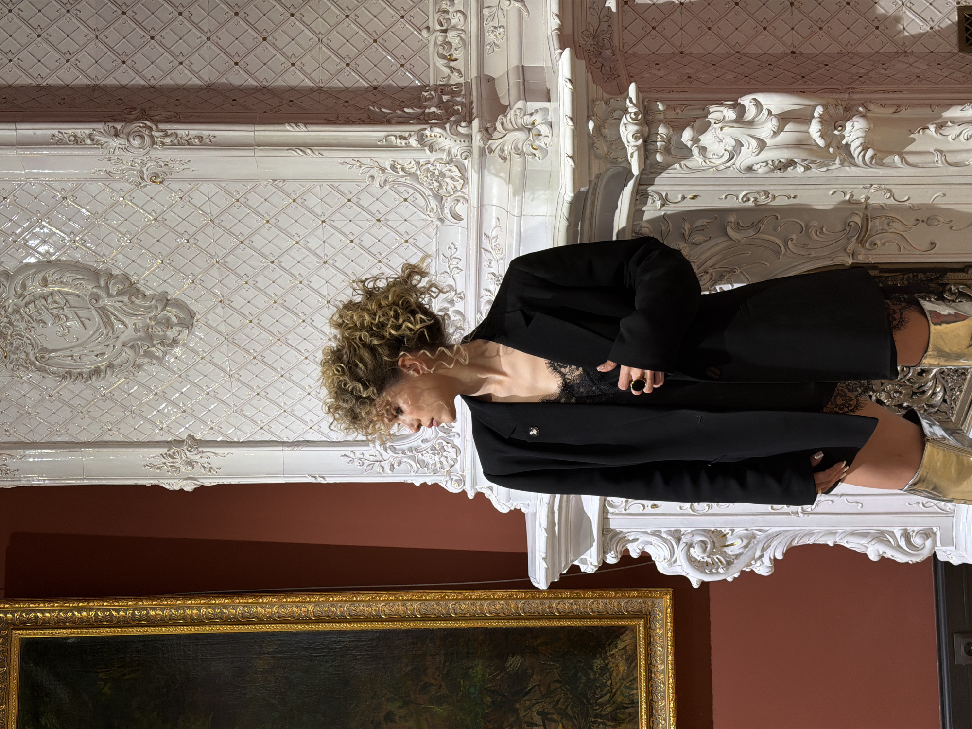 A woman with curly blonde hair is the subject of this image, wearing a black blazer over a black lace top. She is positioned near a decorative white fireplace with intricate carvings and tiles. Below the fireplace, a framed painting with a gold frame is visible. The background is a deep reddish-brown wall. The location is likely in Kazan, Russia, based on the style and architectural details of the interior, especially the ornate fireplace.