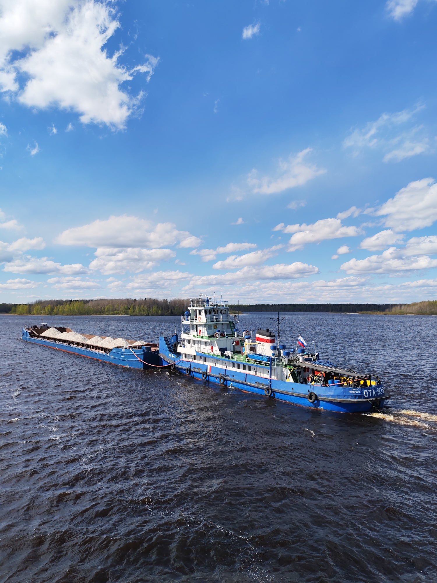 A blue and white tugboat, labeled "OTA-929" and "Санкт-Петербург" (Saint Petersburg) on its hull, is pulling a barge loaded with a light-colored granular material across a body of dark water. The tugboat is moving, as evidenced by the wake behind it. A Russian flag is visible on the tugboat. On the barge, the granular material is heaped into several mounds. The shoreline is visible in the background, with a line of trees. The sky is blue with scattered white clouds. There are no people distinctly visible. The location is not specified as Pochinok, Russia in the image.