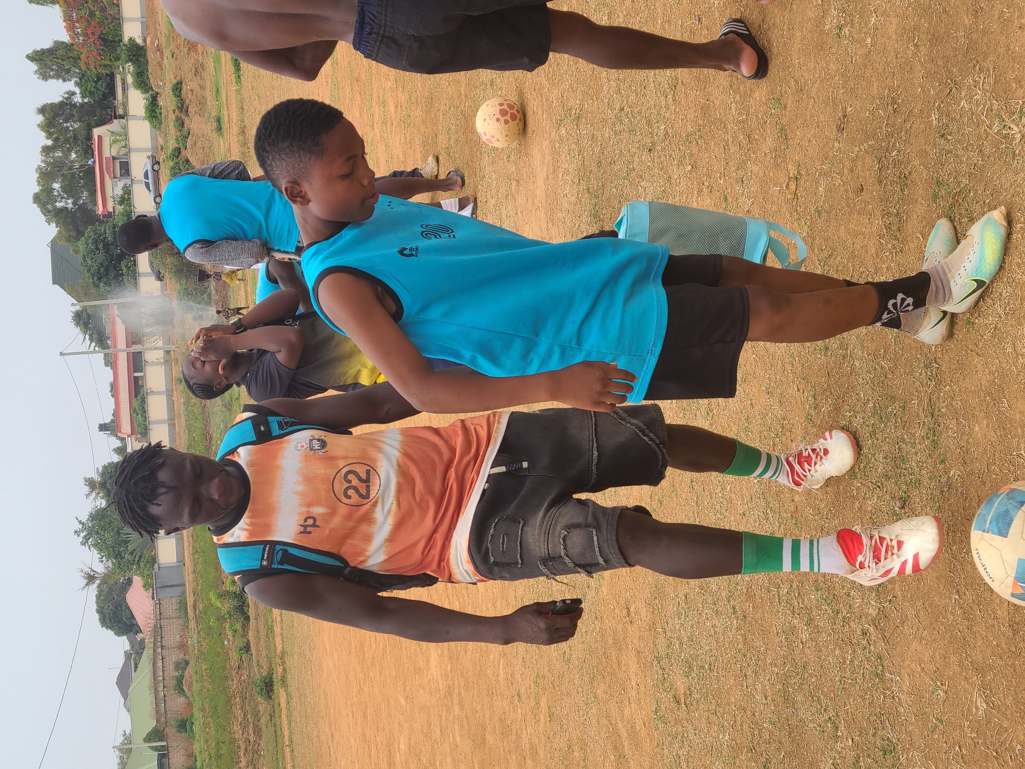 The image depicts an outdoor scene in Barikin Jirga Summa, Nigeria, featuring a group of young people, seemingly engaged in a soccer practice or casual game. Two individuals are prominently positioned in the foreground. One is wearing an orange and white tie-dye patterned shirt with the number "22" on it, paired with black denim shorts and green and white striped socks. He is holding a small black object in his hand. The other person is wearing a bright blue tank top and black shorts.
Several other individuals are visible in the background, some also wearing blue shirts. There are two soccer balls visible in the image. In the background, there are buildings and vegetation, including trees and grass. A plume of steam is visible. The ground appears to be a dry, grassy surface, possibly a field or sports ground.