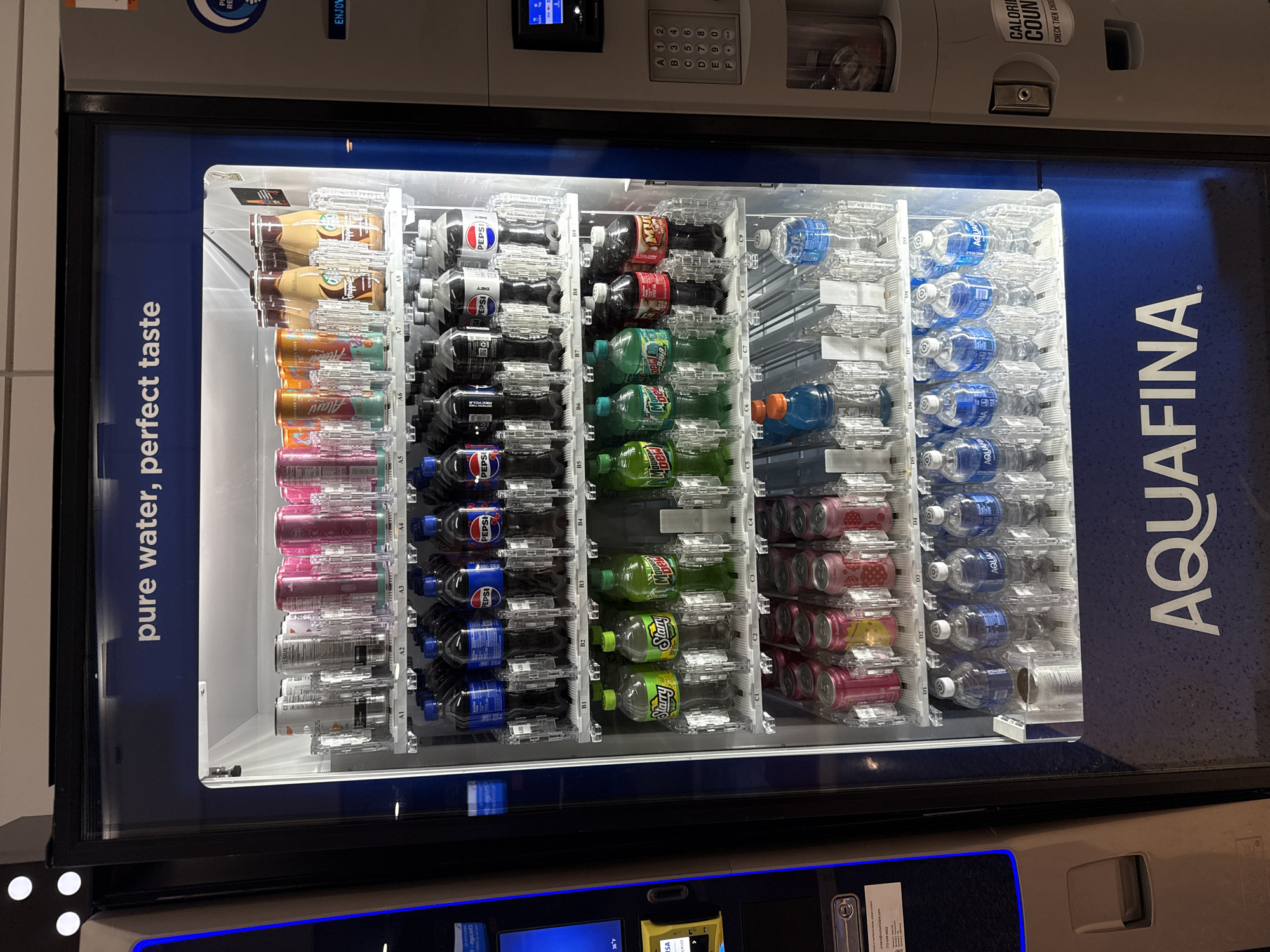 The image shows a well-stocked vending machine filled with various drinks, including Starbucks Frappuccino, Alani Nu energy drinks, Pepsi, Diet Pepsi, Mtn Dew, Starry, Mtn Dew Major Melon, Gatorade, Aquafina water, and various other canned drinks. The vending machine is marked with "Aquafina" branding on the right. The image also shows the numeric keypad and screen for making a purchase. The machine is likely located in Chicago, based on the information provided.
