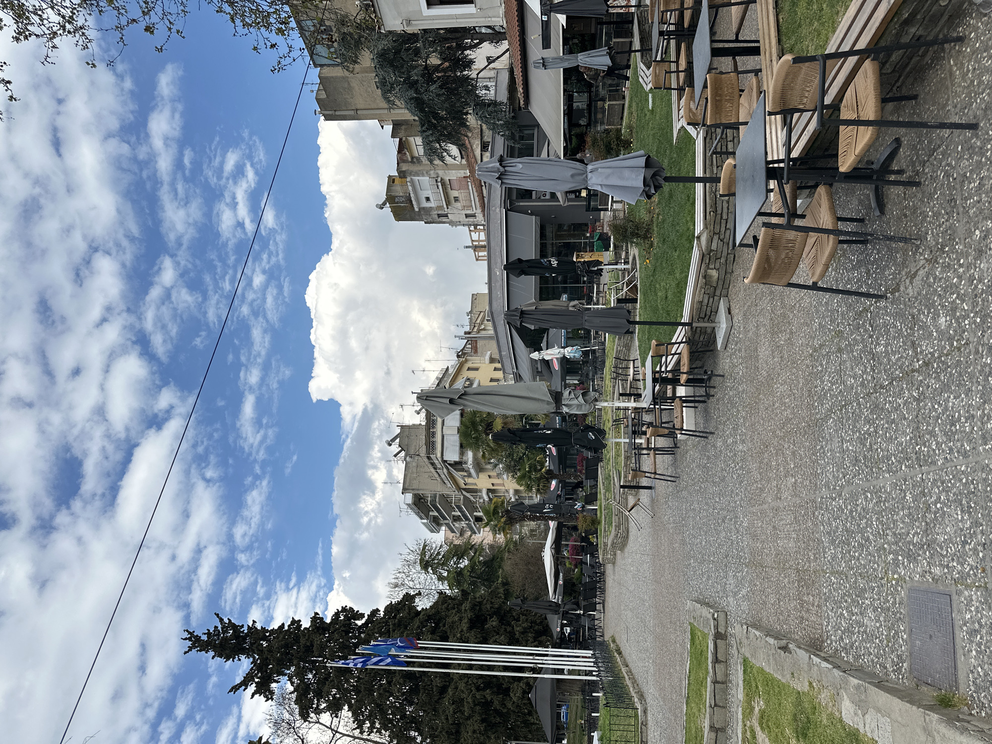 The image captures a street scene in Serres, Greece. A row of Greek flags stands near trees, under a blue sky with clouds. Alongside the flags, a pedestrian walkway leads toward a series of outdoor cafes. The cafes have tables and chairs covered by umbrellas. Buildings line the opposite side of the walkway, adding to the urban setting. The overall atmosphere suggests a quiet day, with the cafes seemingly not busy at the time the photo was taken.