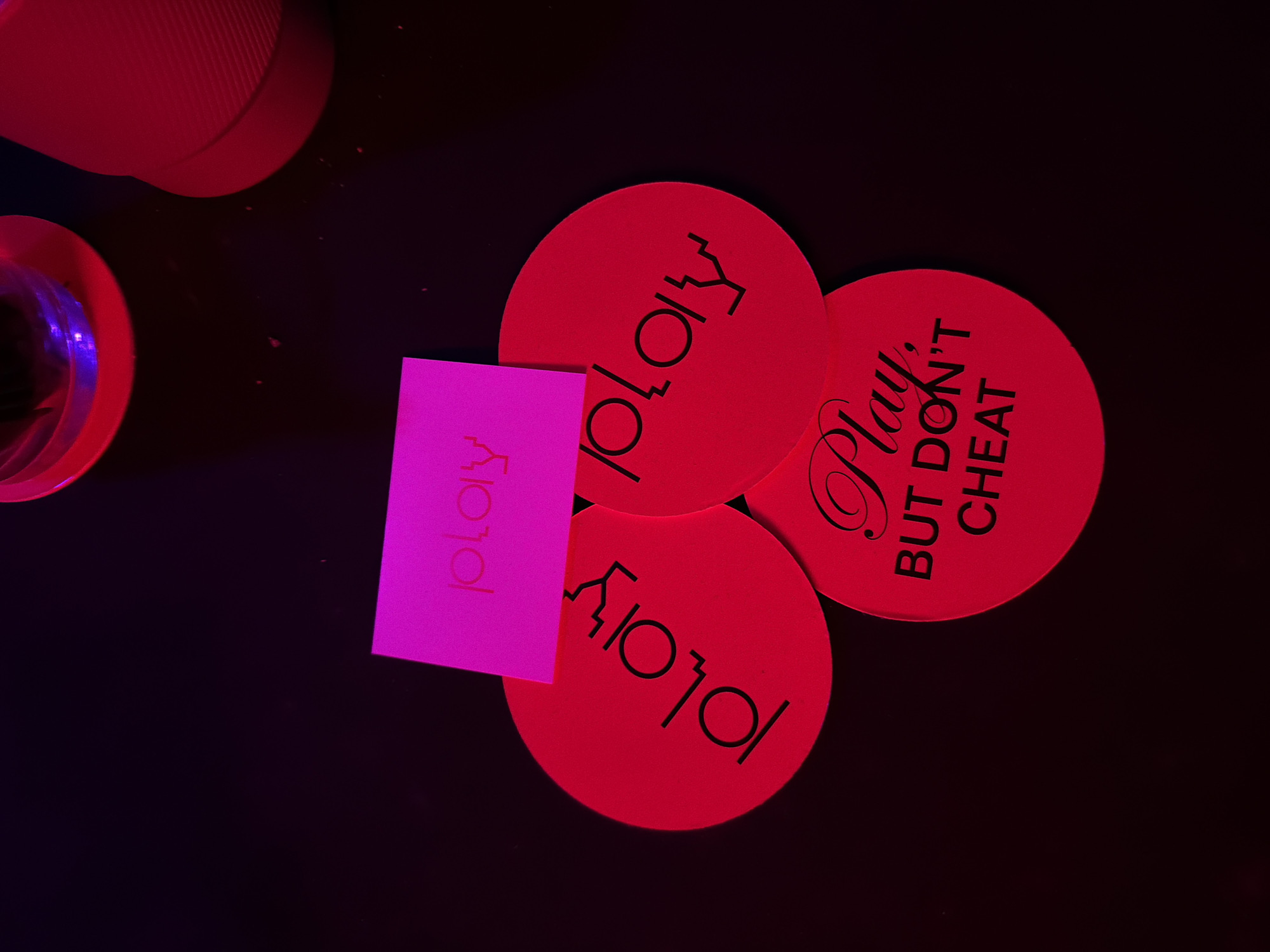 A high-angle shot shows four round, red coasters scattered on a dark surface along with a small pink card. Two of the coasters display the word "PLAY" in black, stylized lettering that is vertically flipped. Another coaster reads "Play BUT DON'T CHEAT" in black lettering. The pink card also has the word "PLAY" printed on it in a similar stylized font but in a smaller size and is flipped vertically. In the upper left corner, portions of a clear glass and a ribbed container are visible, both illuminated with a pinkish-red light. The location is likely a bar in the city of Sofia, Bulgaria.