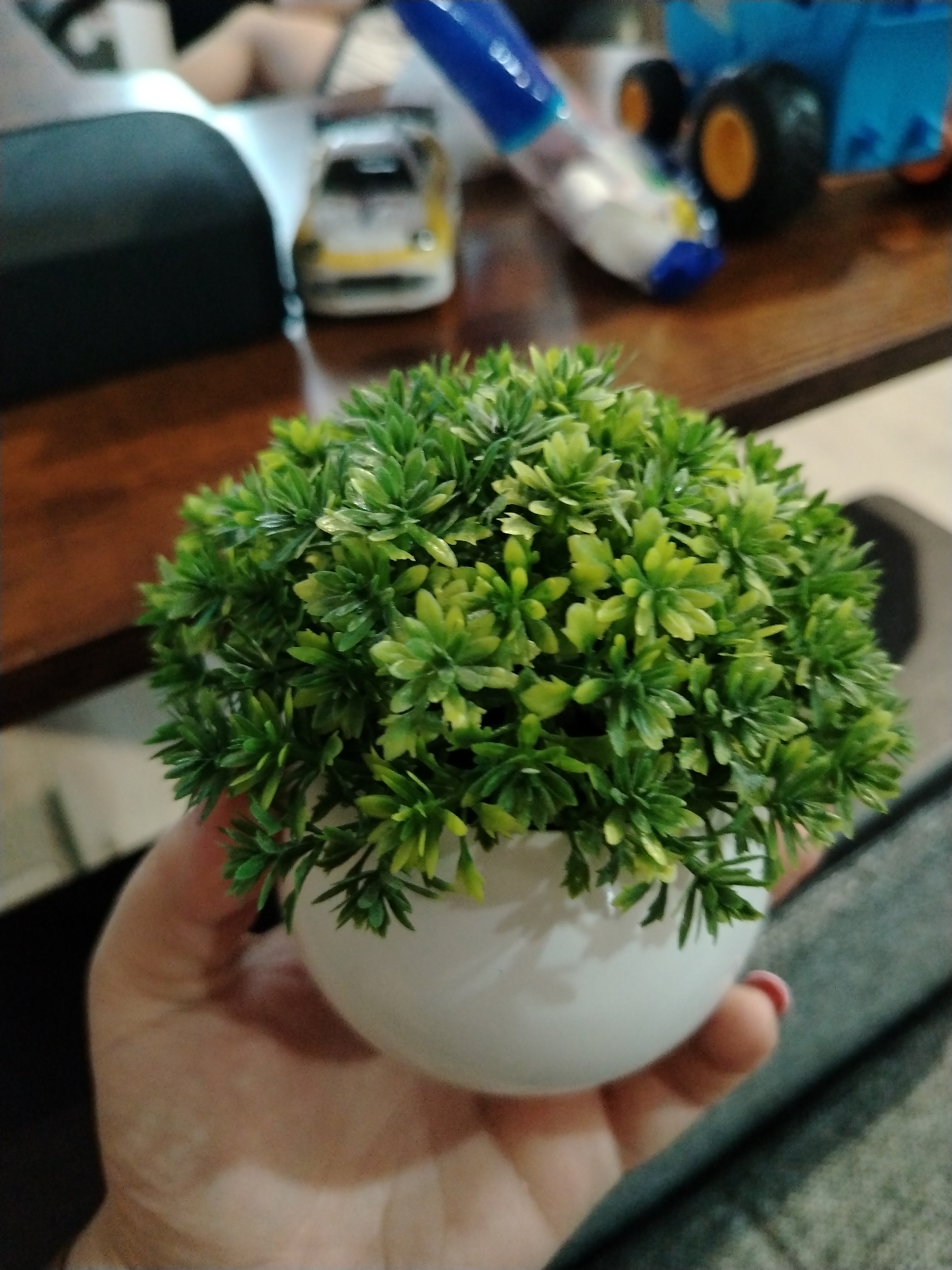 An indoor close-up shot features a person's hand holding a small, artificial plant in a glossy white spherical pot. The plant consists of dense, spiky foliage in vibrant shades of green and light green, arranged to form a compact, rounded shape. The hand holding the pot has light skin, and a hint of pink nail polish is visible on one finger.
The background is significantly blurred, indicating a shallow depth of field that keeps the plant in sharp focus. Various objects are subtly visible in the soft-focus background on a wooden surface. To the left, a dark-colored object, possibly a cushion or a piece of furniture, is partially seen next to what appears to be a person's legs or feet. Further back and to the left, a blurred yellow toy car with white accents is discernible. On the right side of the background, a blue and orange toy truck, likely a dump truck, with black and yellow wheels, is visible. An object resembling a marker or pen with a blue cap can also be spotted. The setting appears to be a casual home environment, suggesting a living room or bedroom. The lighting is consistent with an indoor daytime scene. There is no visible text in the image, and no specific emotions are expressed.
