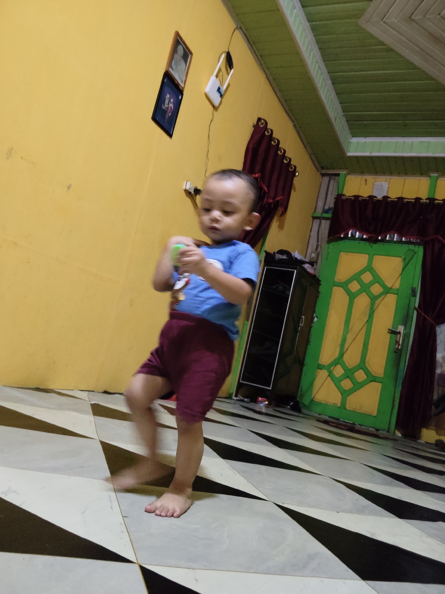 In a room with yellow walls and a green and white patterned ceiling, a toddler wearing a blue t-shirt and maroon shorts is captured mid-stride on a black and white tiled floor. The child is holding a small, light green object. To the right of the child, a tall, dark cabinet stands against the wall, which is adorned with burgundy curtains. Beyond the cabinet, a bright green door with yellow geometric patterns is visible. On the yellow wall to the left, two framed pictures hang. The scene appears to be indoors, likely a residence, in Tanah Grogot, Indonesia.