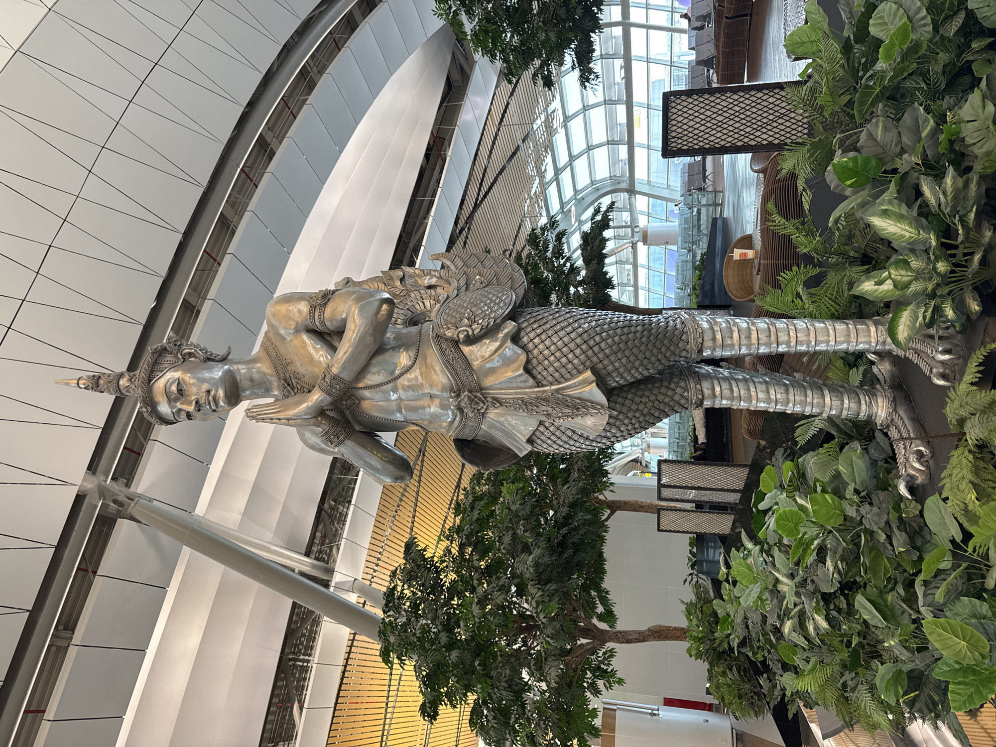 The photograph shows a silver statue of a Thai mythological figure, likely a Kinnaree, in an indoor setting. The statue is surrounded by greenery. It is positioned in an atrium, with a curved, multi-paneled ceiling and large windows allowing natural light. The location is likely a commercial or public building in Nong Prue Subdistrict, Thailand. The surrounding environment features modern architectural elements and interior design, including seating areas visible through the windows.