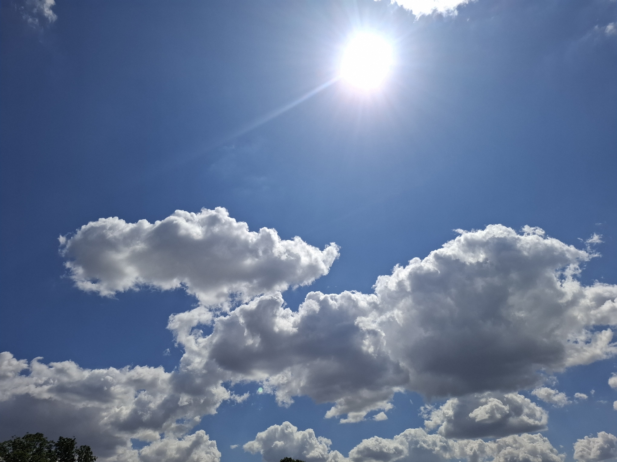 A bright sun shines in a clear blue sky dotted with fluffy white clouds. The sun is located in the upper right quadrant of the image, casting a bright light with visible rays. Several large cumulus clouds are scattered across the sky, with some appearing darker and more ominous towards the bottom. The bottom edge of the image shows the tops of green trees, suggesting an outdoor scene in a relatively natural or park-like setting. There are no people or specific man-made objects visible in the image besides the implied presence of the sky and clouds, which are natural elements. The scene depicts a sunny day.