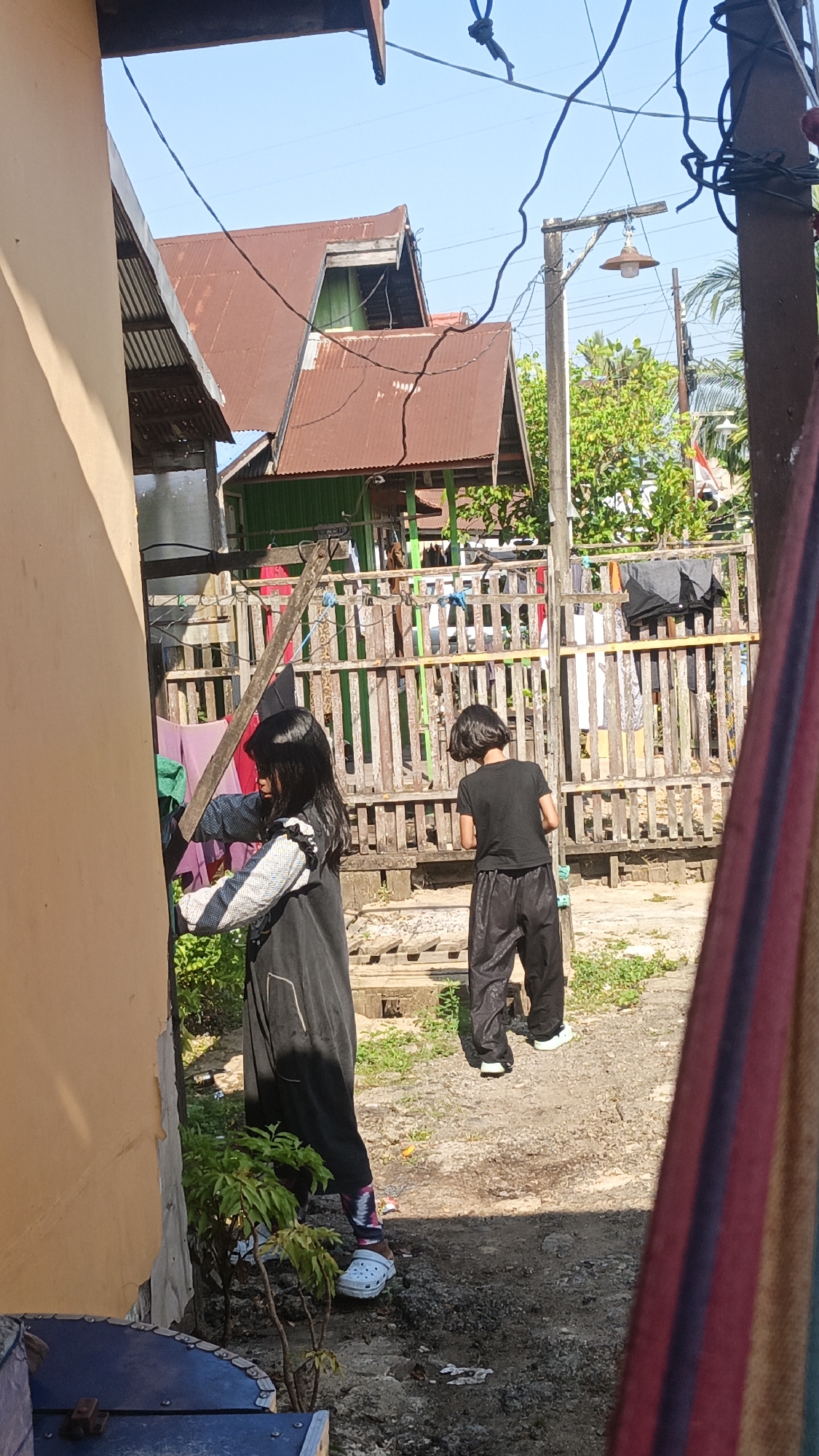 The image depicts a sunny outdoor scene, likely in a residential area of Banjarmasin, Indonesia, characterized by a rustic wooden fence and several residential structures with corrugated metal roofs. In the foreground, a young girl with long dark hair is partially visible on the left, wearing a dark jumpsuit with a patterned sleeve. She appears to be interacting with a hanging object, possibly laundry. To her right, another young person, also with dark hair, is standing further back, wearing a black t-shirt and loose-fitting dark pants. This individual is facing away from the camera and appears to be observing something on the ground. Clothes are hung on the wooden fence, suggesting a laundry area. The ground is a mixture of dirt and gravel, with some sparse vegetation. A wooden utility pole with a hanging lamp is visible in the mid-ground. The overall impression is a casual, everyday scene in a tropical or subtropical environment.