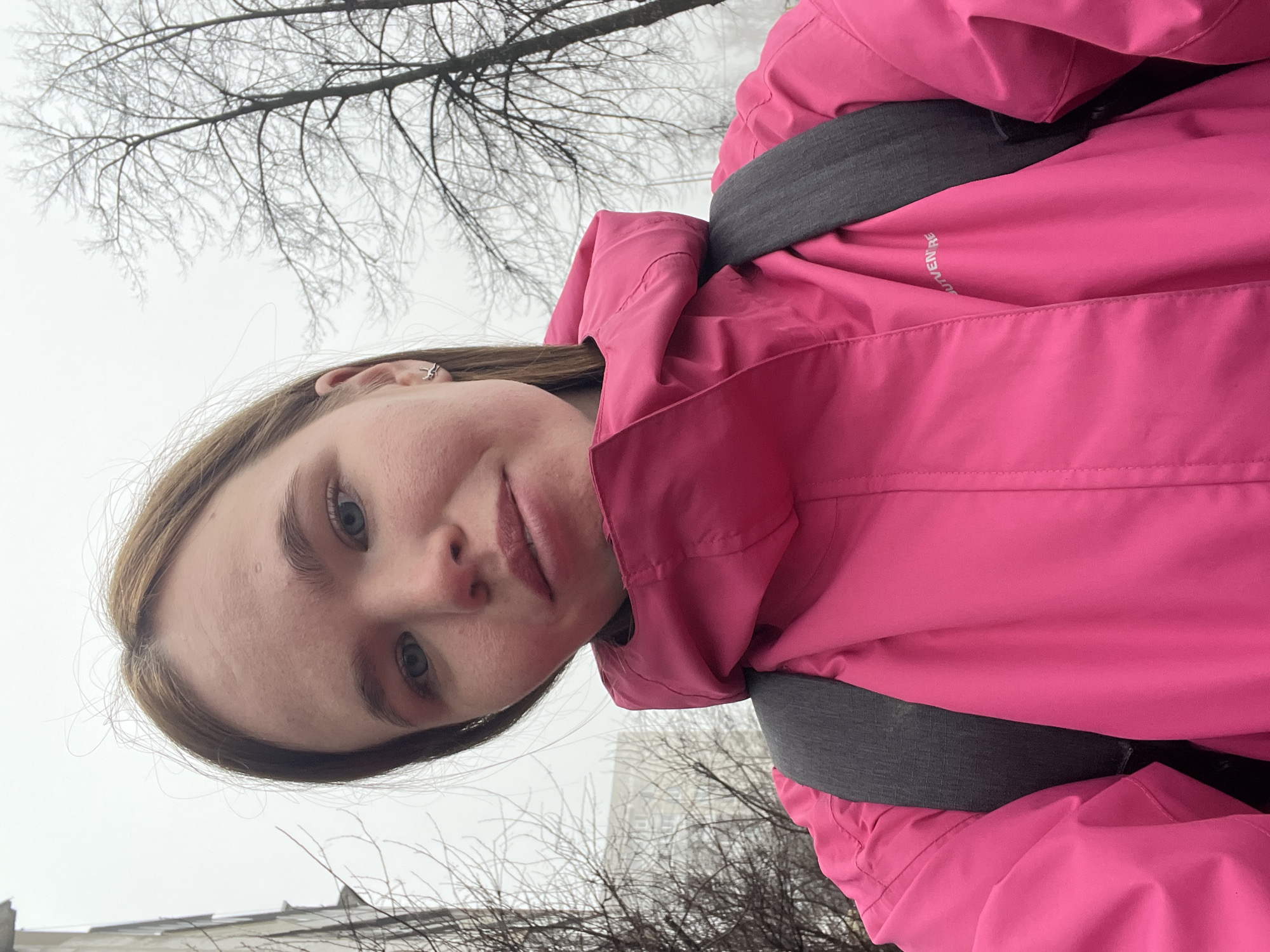 A young woman is depicted in a selfie-style photograph taken outdoors on an overcast day. She is wearing a bright pink hooded jacket with gray backpack straps visible. The background includes bare tree branches and a gray, overcast sky. The location appears to be urban, given the presence of buildings and trees. The photograph was taken in the city of Chaikovsky, Russia.