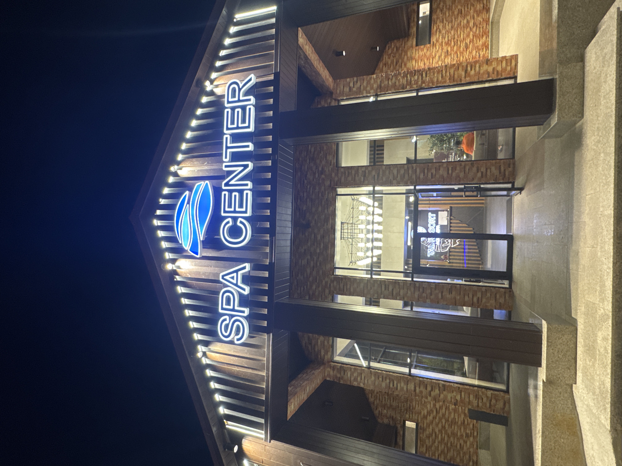 The image shows the exterior of a "SPA CENTER" building at night, located in Talgar, Kazakhstan. The building has a prominent neon sign with the words "SPA CENTER" in blue and white, with a blue wave-like logo above it. The facade is constructed with wooden planks and brickwork. A glass entrance leads into the building where a reception desk with the text "TAIL RESORT" is visible. Inside, a person wearing an orange shirt is seated on a sofa. The area in front of the building is paved and lit.