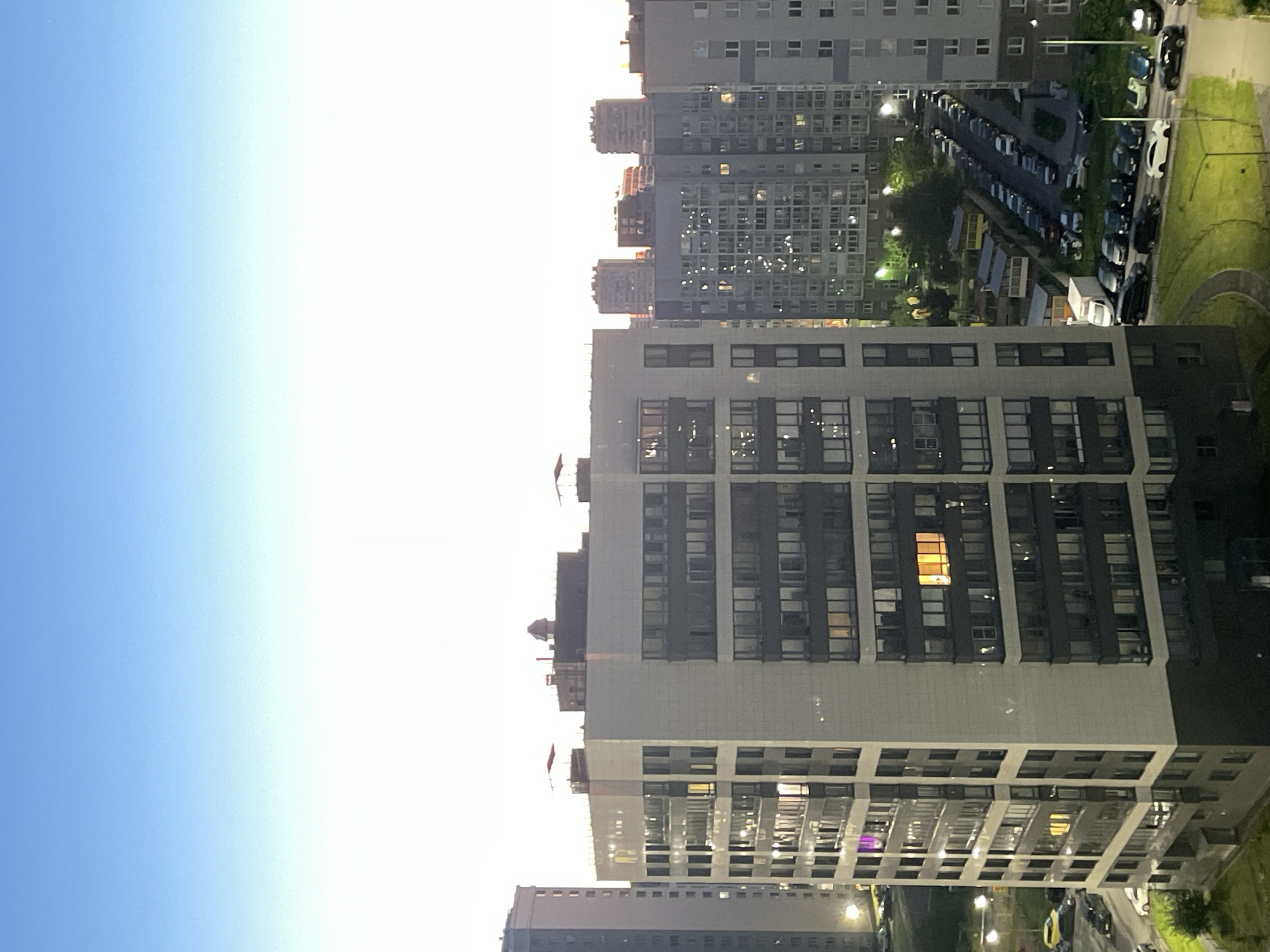 The image shows a high-angle view of a residential area in Sinyavino, Russia, during twilight. A prominent, modern, multi-story apartment building with a light-colored facade and dark window frames dominates the foreground. Multiple windows in this building are illuminated, suggesting occupancy. Adjacent to this building, further into the cityscape, are other apartment buildings of varying heights and architectural styles. Some of these buildings also have illuminated windows, creating a speckled pattern against the darkening sky. In the background, the sky transitions from a clear light blue at the top to a bright, almost white horizon near the buildings, indicative of dusk. The lower portion of the image reveals a street lined with parked cars, primarily dark-colored sedans and SUVs, as well as some white vehicles. Streetlights are visible, casting pools of light on the pavement and surrounding grass. A small green area with a curved pathway is also visible in the lower right quadrant. There are no people clearly visible in the scene. The overall impression is of a typical urban evening scene in a Russian residential district.
