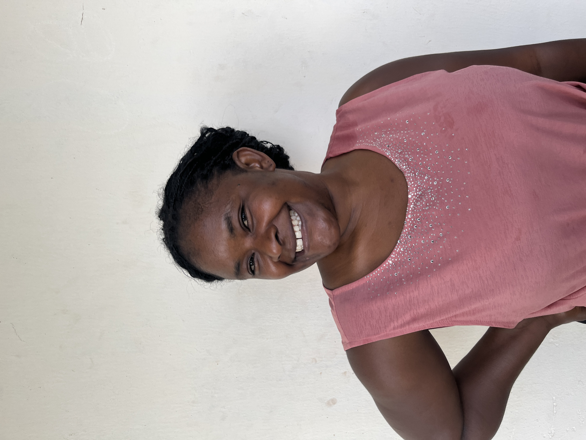 A medium shot portrait of a Black woman with braided hair, standing in front of a textured, off-white wall. She is smiling broadly and looking towards the viewer. Her skin is dark brown, and her teeth are visible in her smile. She is wearing a sleeveless pink top adorned with small, iridescent embellishments around the neckline. Her left arm is bent, with her hand resting on her hip. The background is a plain, slightly mottled wall, indicating an indoor or sheltered outdoor setting, possibly in Gura Top, Nigeria.