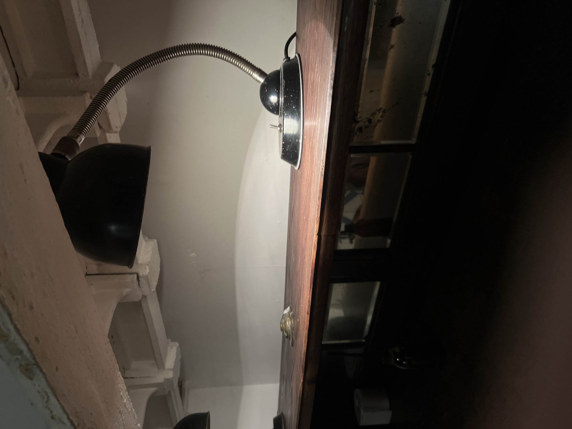 A black metal desk lamp with a flexible gooseneck is positioned on a dark wooden countertop. The lamp's base is round and black with white speckles, featuring a silver toggle switch. A black power cord extends from the base. The countertop itself appears to be part of a counter or shelf with dark paneled sections beneath it, some of which have reflective surfaces or glass. To the left of the lamp, there is a small, reflective object on the countertop, possibly a coin or button. Further to the left, a roll of white paper is visible. The background consists of a plain, light-colored wall. The lighting suggests an indoor environment, likely dimly lit, with the lamp being the primary light source in the immediate vicinity. No people are present in the image. The context of Nizhny Novgorod, Russia, is not discernible from the visual information provided in the image.
