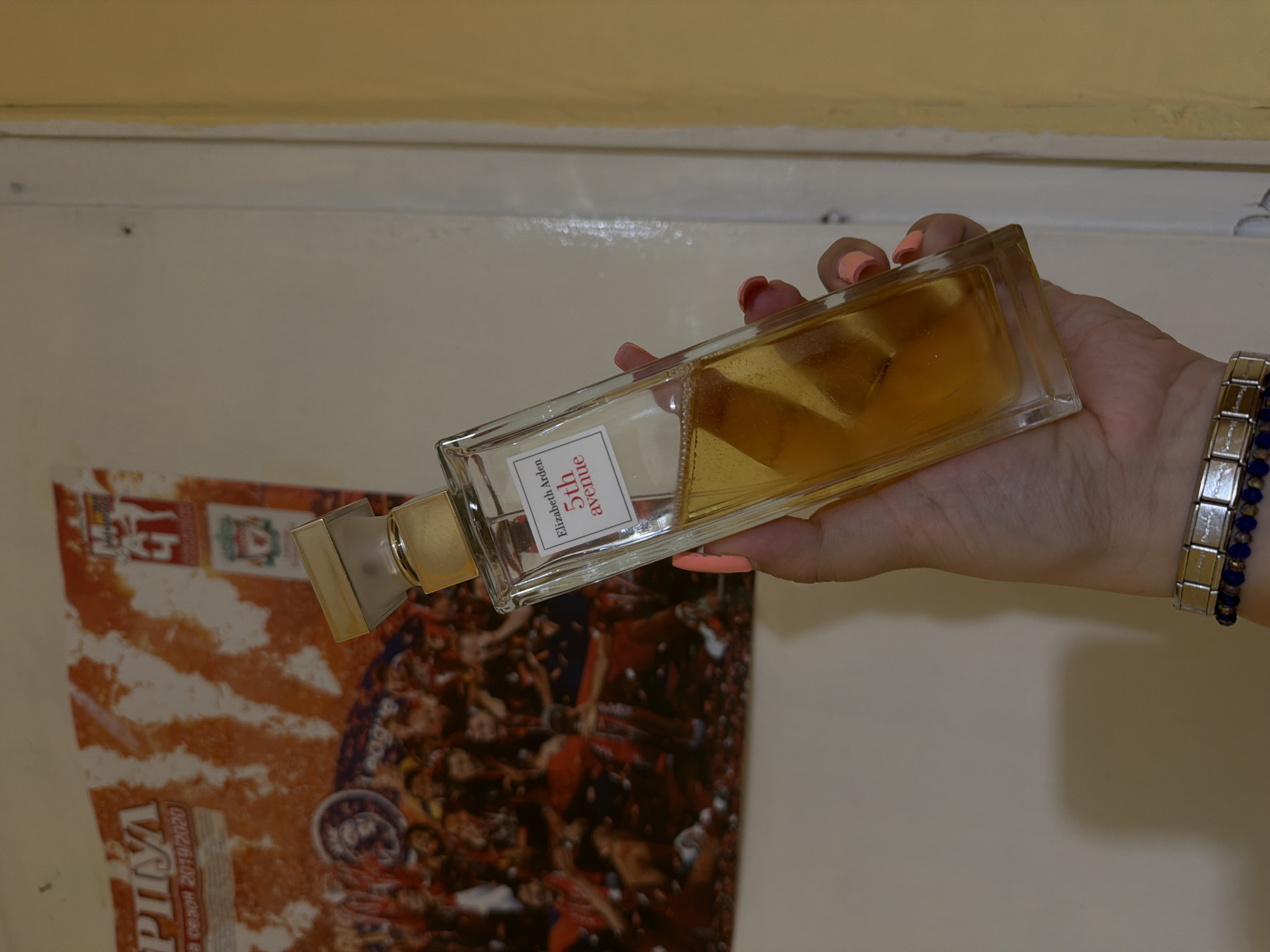 A hand holds a bottle of Elizabeth Arden 5th Avenue perfume against a background consisting of a wall and a poster. The hand is adorned with a bracelet, and the person has manicured fingernails. The perfume bottle is partially full with a yellow-hued liquid. The poster shows a collage of figures with orange and red coloring, along with a seal or emblem near the bottom, and the words "PIVA" and "CE3OH 2019/2020". The photograph's location is Senovo, Bulgaria, but the image itself does not provide specific details or landmarks that pinpoint a more exact location within the city.