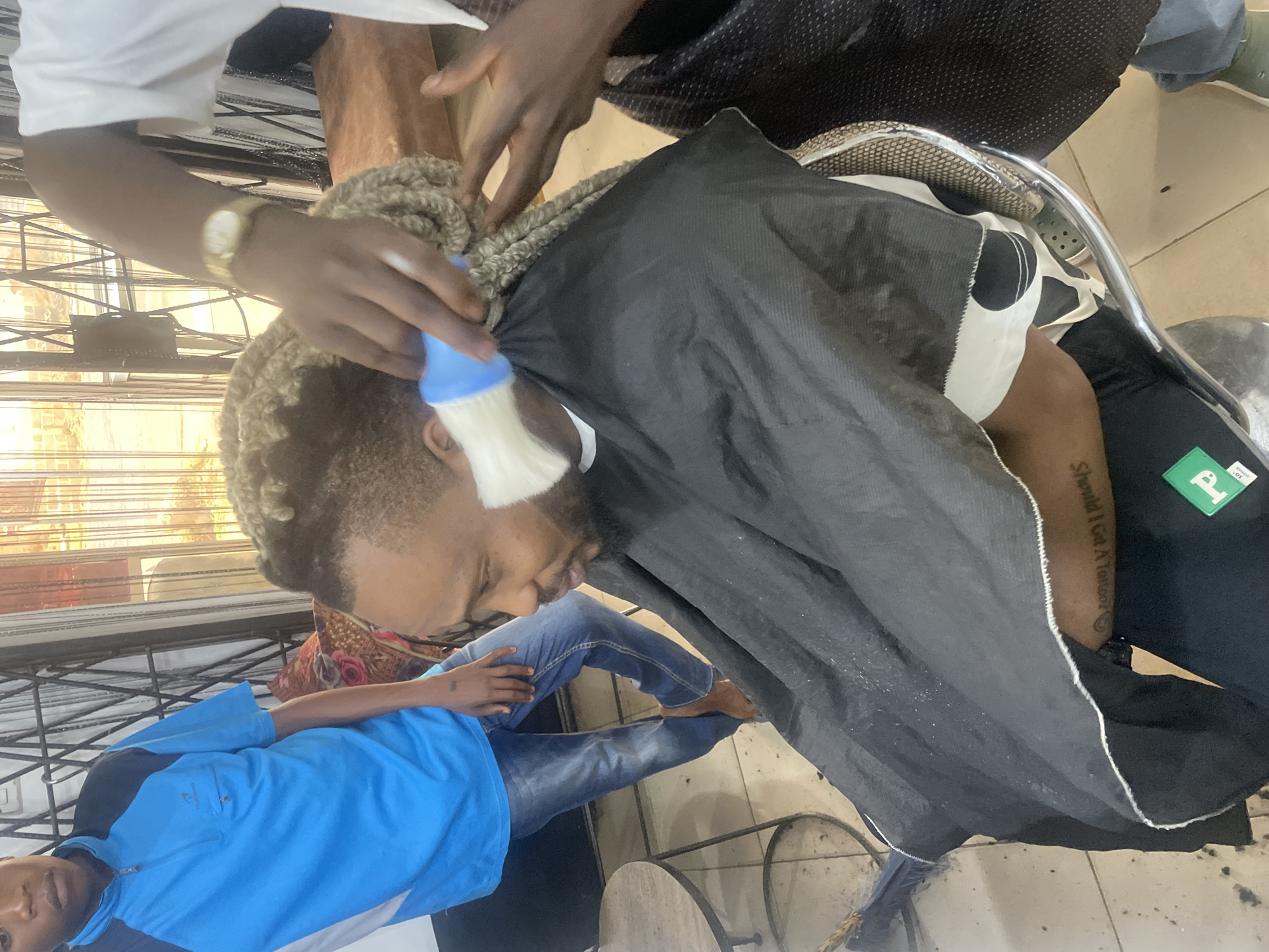 A man is getting a haircut at a barbershop in Jos, Nigeria. He is sitting in a barber chair with a black cape draped around him. The barber is using a brush to remove hair from his head. The man has short, dyed blonde hair. Another person, presumably a helper, is seated off to the side.