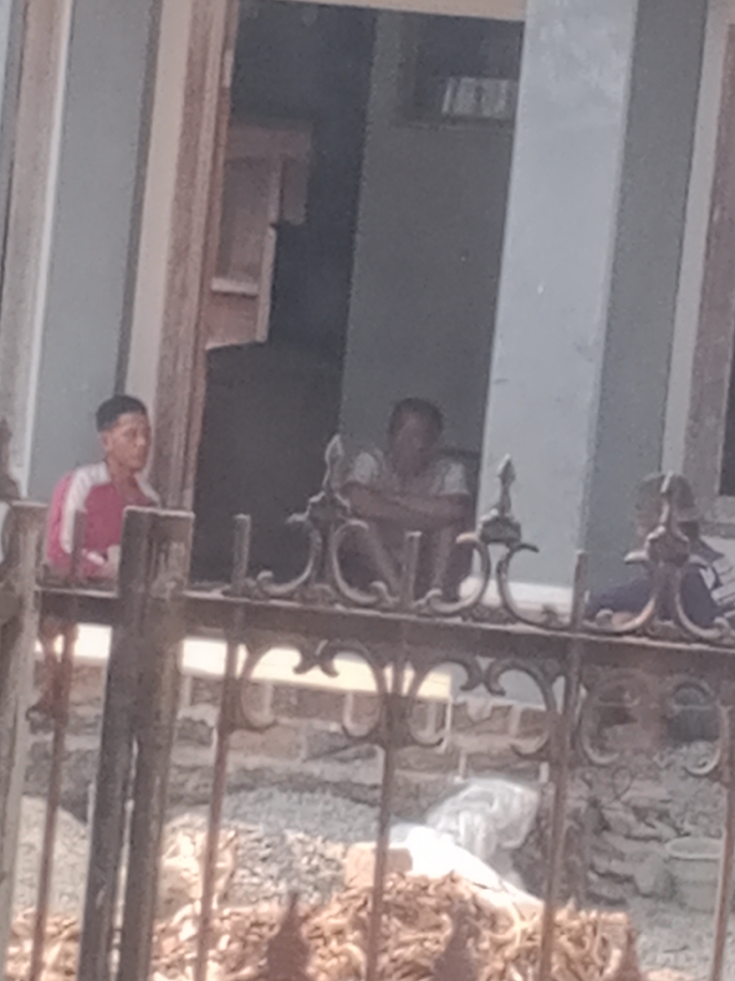 This is a medium-shot, slightly blurry photograph taken outdoors, likely in a residential area. In the foreground, a dark, decorative metal fence runs horizontally across the frame. Behind the fence, a young man wearing a red and white striped shirt stands leaning on what appears to be a railing or balcony. He is looking to his right, away from the camera. Further to the right, two individuals are visible sitting down, partially obscured by the fence and the low resolution of the image. One is a man with his arms crossed, and the other, to his right, appears to be wearing a dark top with lighter markings. The background shows part of a building with an open doorway or window, revealing a darker interior space. The ground behind the fence is covered in loose gravel and some debris, including what looks like white plastic sheeting and piles of natural materials, possibly for construction or gardening. The lighting suggests it is daytime, with soft light illuminating the scene. The location is identified as Lembah, Indonesia.