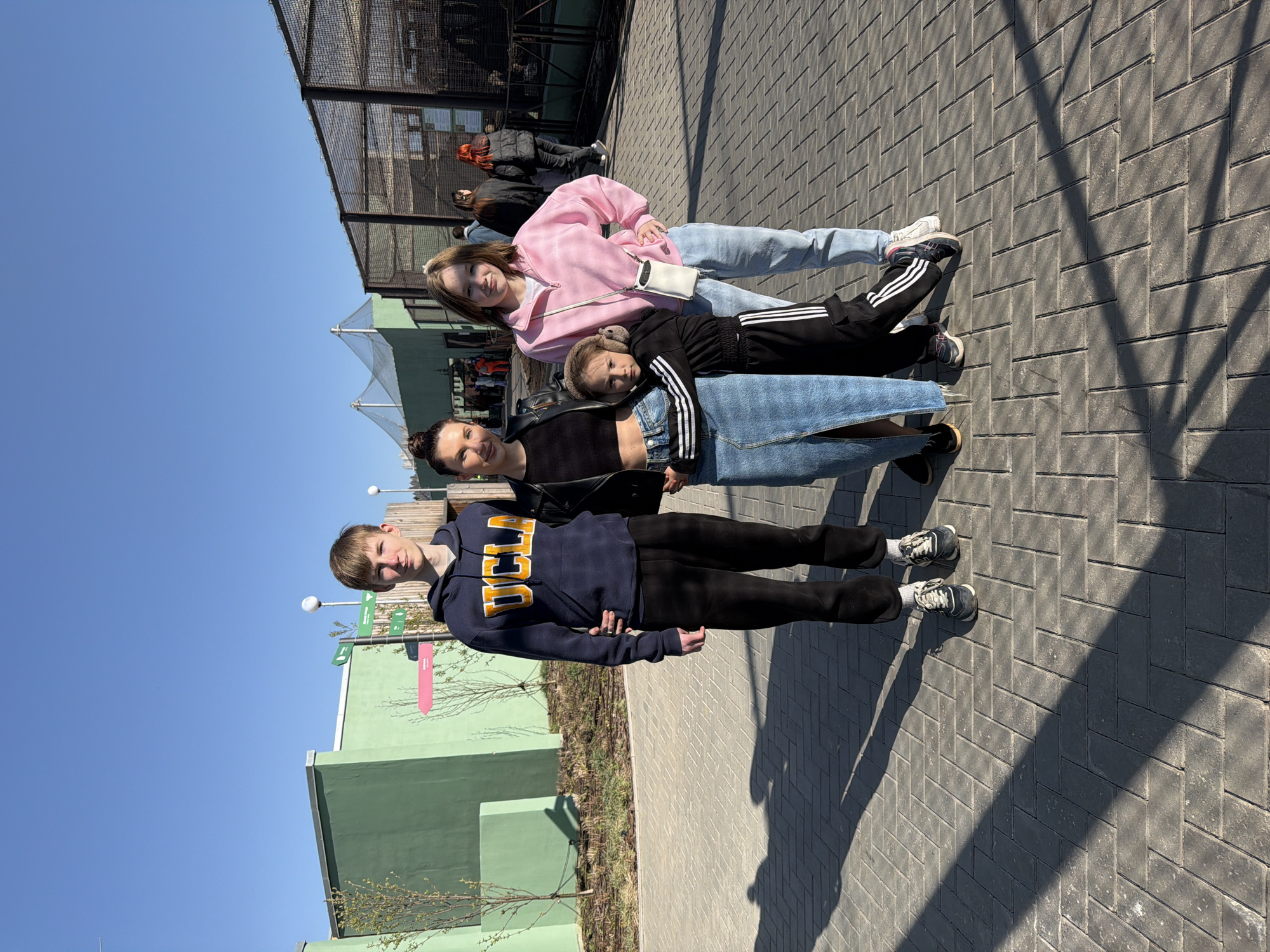 A group of four individuals are positioned in a vertical line on a brick sidewalk, seemingly posing for a photograph. The group consists of what appear to be a mother with three children of varying ages. The individual at the bottom of the line is a teenage boy wearing a navy blue hoodie with the letters "UCLA" in gold and black pants. The woman in the middle wears a black top, a black leather jacket, and a denim skirt. On top of her is a young child wearing a black tracksuit with white stripes. The individual at the top is a teenage girl in a pink hoodie and light blue jeans. The background includes a green building and a blue sky on the left, and a metal fence and other people in the distance on the right. The location is Perm, Russia.