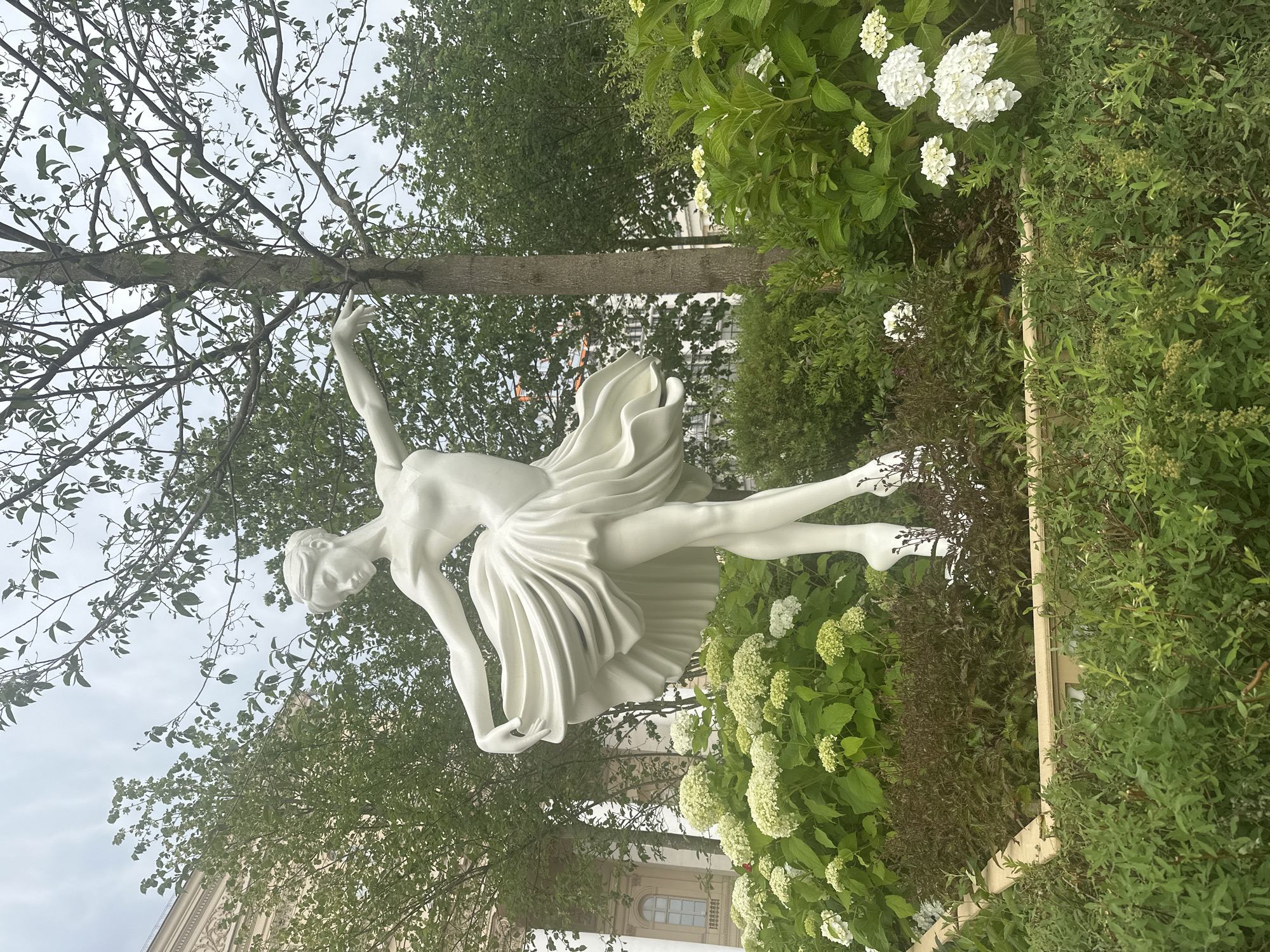 This is a vertical, eye-level, full shot photograph of a white statue of a ballerina in a garden. The statue is positioned on the right side of the frame, in mid-pose with one arm extended upwards and the other bent, her body arched. She wears a pleated skirt, and her hair is pulled back.
The ballerina statue is surrounded by lush greenery, including dense bushes with small white flowers, likely hydrangeas, and the branches of a large tree with sparse leaves. The tree trunk runs horizontally across the middle of the image. In the background, a building with windows is partially visible through the foliage. The sky is overcast and cloudy.
The scene appears to be set outdoors in a park or a garden. The lighting suggests it could be daytime, possibly overcast weather given the sky. The overall mood is serene and artistic, highlighting the graceful form of the statue against the natural backdrop. The location is identified as Moscow, Russia, though no specific landmarks are clearly identifiable. There is no visible text in the image.