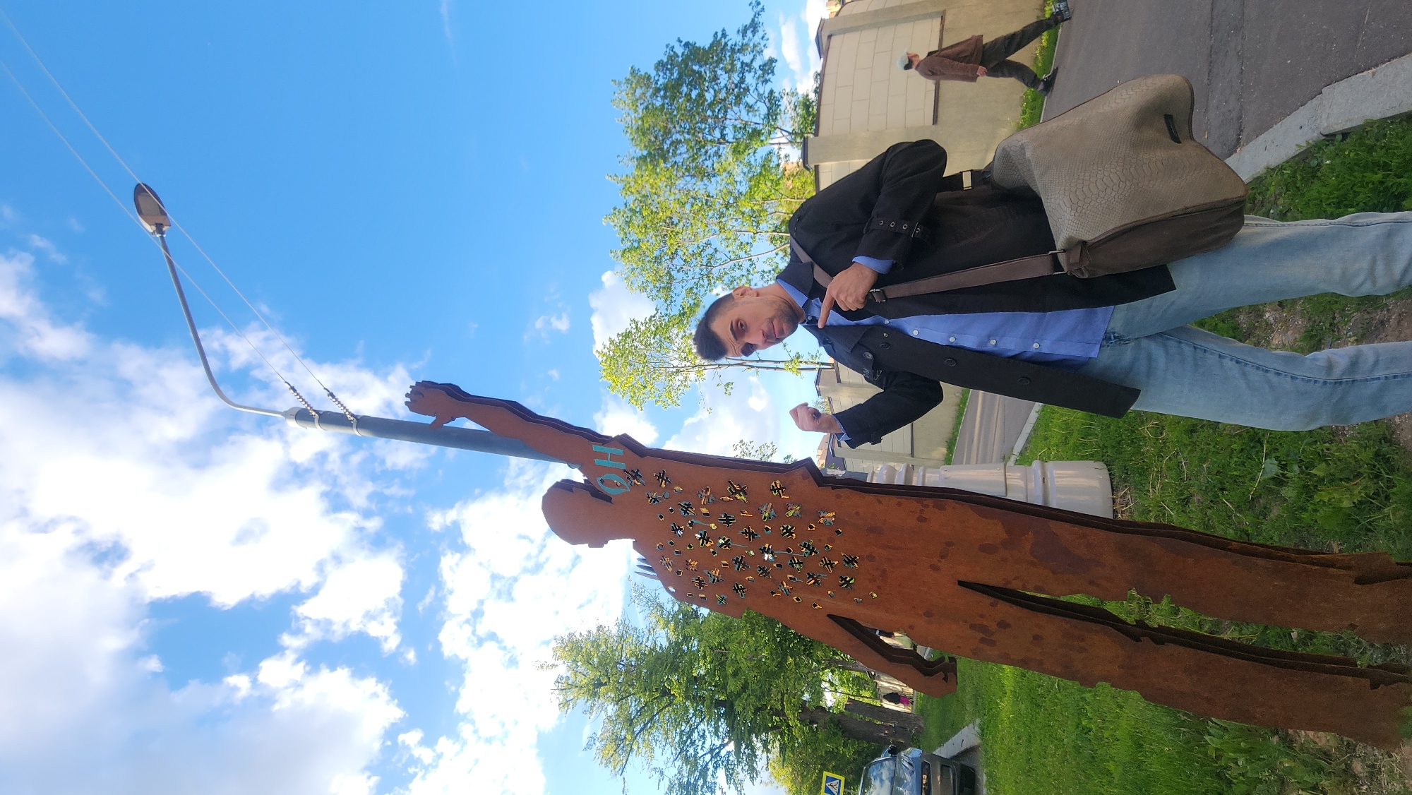 A man poses with a metal sculpture in Moscow, Russia. The sculpture, depicting a human figure, is made of rusted metal with numerous cross-shaped cutouts across its torso. The man, wearing a blue shirt, black coat, and jeans, stands to the right of the sculpture, gesturing toward it. He carries a shoulder bag. In the background, a street lamp rises, and trees with green leaves dot the landscape. A second person walks along the sidewalk in the upper-right corner of the frame. A car is partially visible at the bottom of the frame. The sky is blue with scattered white clouds.