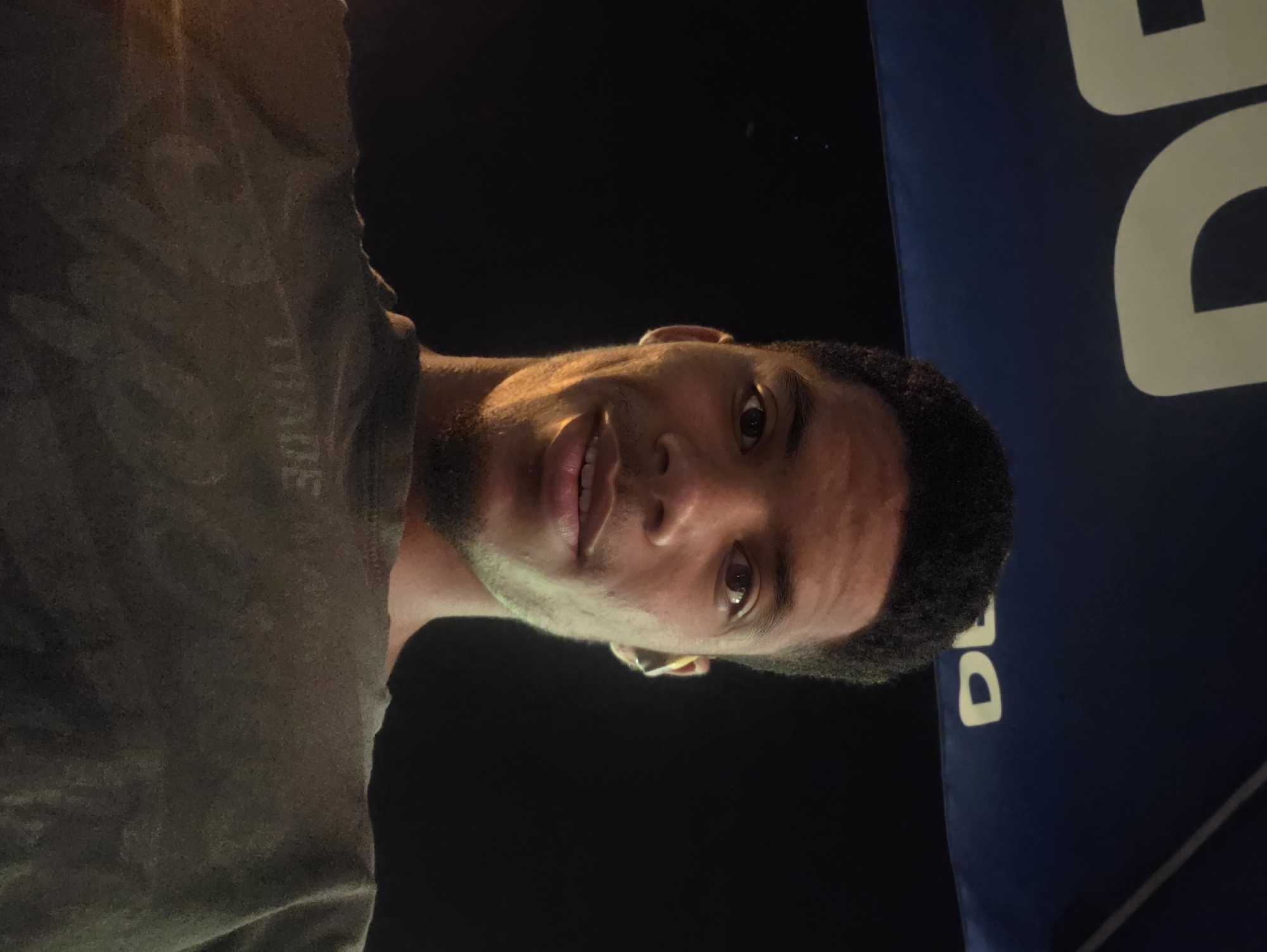 A close-up, low-angle selfie of a young Black man looking directly at the camera. He has short, dark, curly hair and is wearing a dark grey t-shirt. A glimpse of a blue umbrella with white lettering, possibly "DE", is visible at the top of the frame. The background is dark, suggesting it is nighttime. The lighting casts shadows across the man's face, highlighting his cheekbones and the left side of his face. An earbud is visible in his right ear. The overall scene appears to be outdoors, possibly in Varna, Bulgaria, given the context provided.