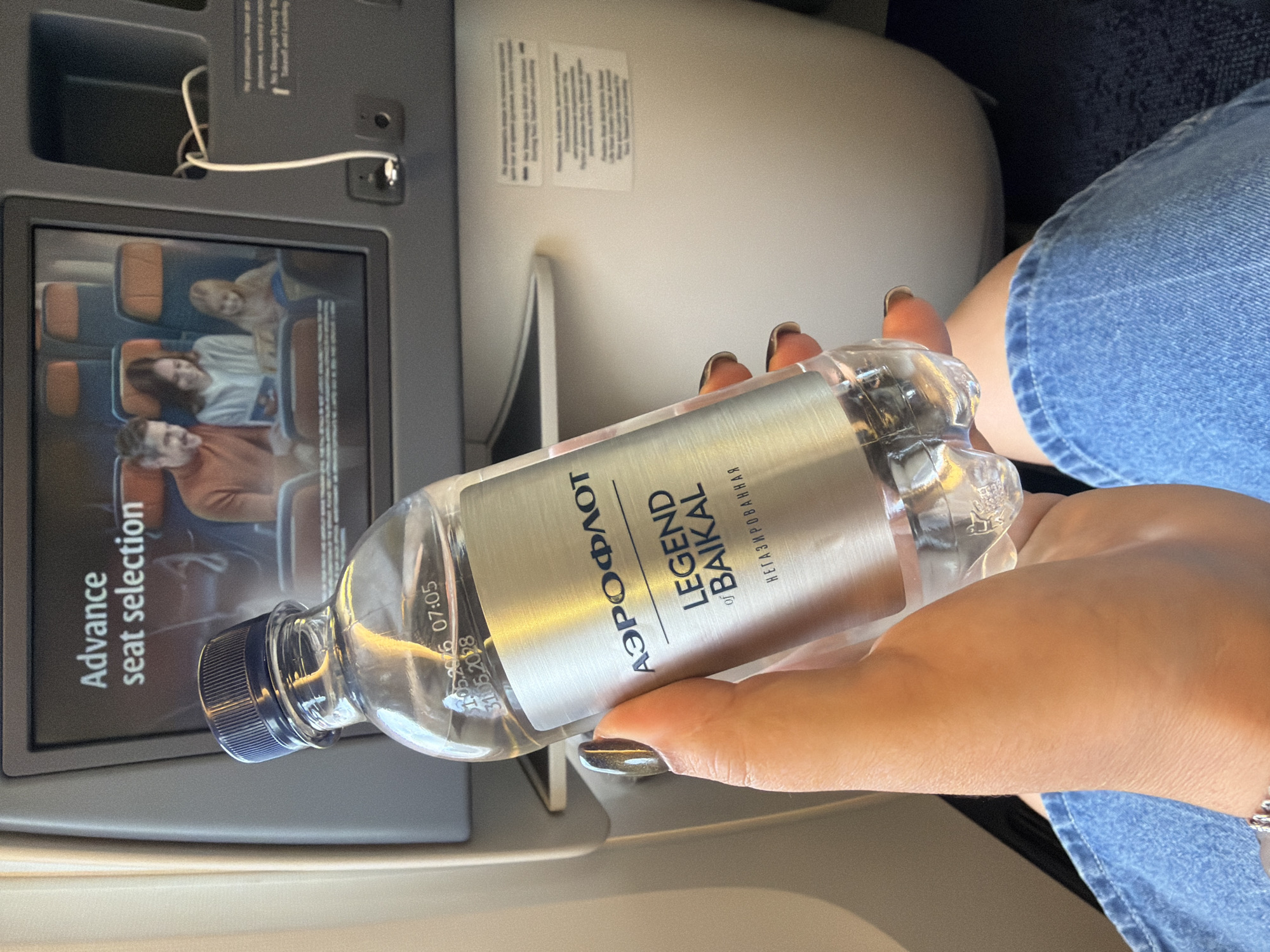A person's hand, with polished nails, holds a clear plastic water bottle with a silver label that reads "АЭРОФЛОТ LEGEND of BAIKAL" and "НЕГАЗИРОВАННАЯ". The bottle has a blue cap. In the background, an airplane's in-flight entertainment screen displays an advertisement for "Advance seat selection" showing a family sitting in airplane seats. The environment appears to be inside an airplane cabin, with visible seating and parts of the cabin's interior. There is a USB port visible on the seatback in front of the screen.