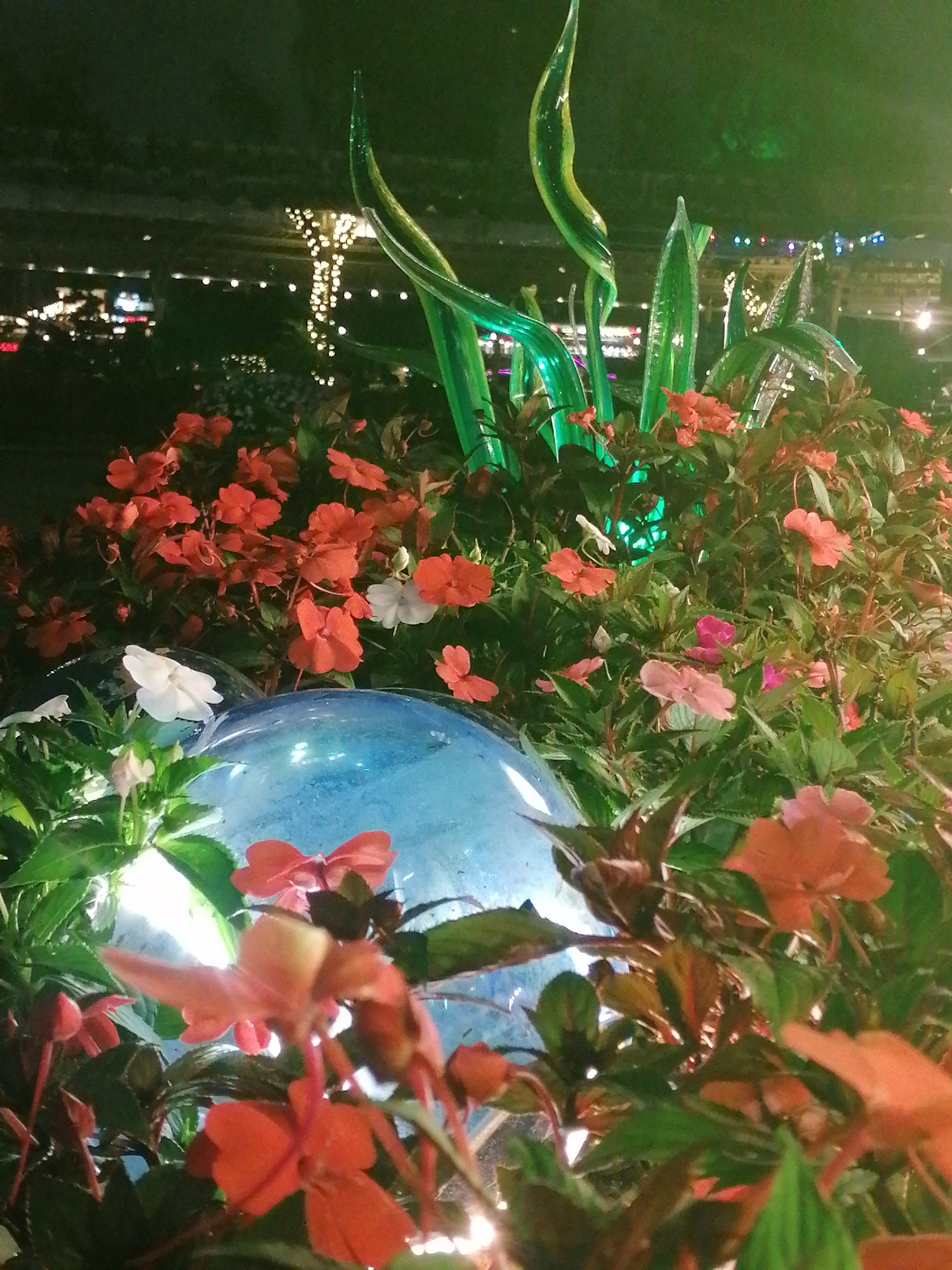 This nocturnal image showcases a beautifully illuminated garden display, likely in a public park or resort in Da Lat, Vietnam. The foreground is dominated by a lush bed of flowering plants, primarily featuring blossoms in various shades of orange and red, interspersed with some white and pink flowers. Their green foliage creates a rich texture.
Rising from the plants are several striking, tall, green, translucent sculptures, which appear to be made of glass or a similar material, designed to resemble stylized plant leaves or stalks. Many of these sculptures emit a soft green glow. Below them, two prominent, spherical blue glass orbs are visible, also radiating light, adding a cool, luminous element to the scene.
The background is dark and out of focus, containing numerous blurred lights, creating a bokeh effect. These distant lights likely belong to buildings, streetlights, or other decorative illuminations, confirming the nighttime setting. The overall lighting scheme gives the garden a magical and ethereal ambiance. No people or visible text are present in the frame. The scene captures a moment of serene beauty and intricate garden design, characteristic of a well-maintained decorative display.