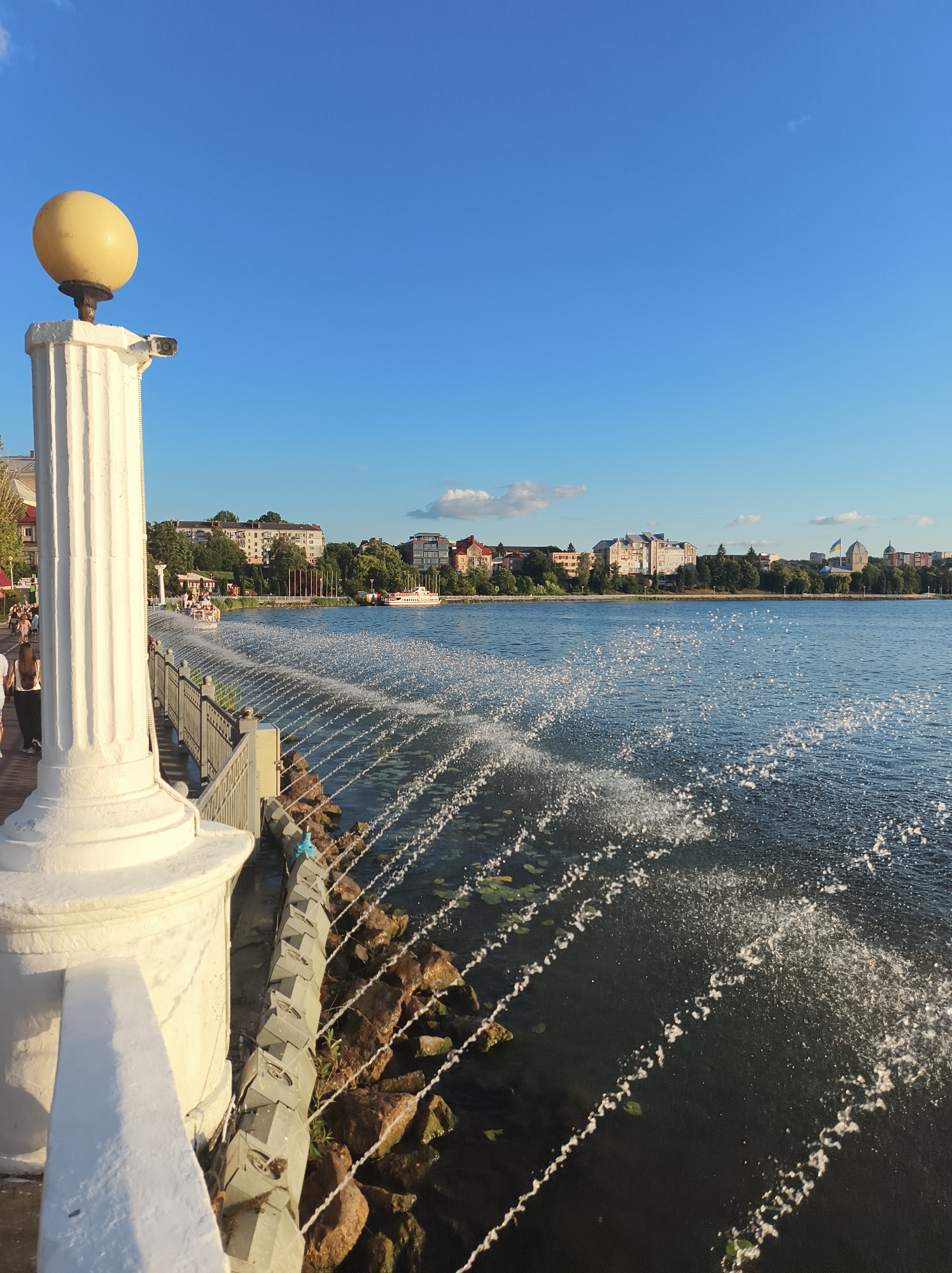 The image captures a sunny afternoon scene at Ternopil Lake in Ternopil, Ukraine. In the foreground, a large white stone column with a yellow lamp on top stands to the left, casting a shadow. To the right, a fountain sprays water in a series of arcs across the dark blue surface of the lake. The water reflects the sunlight, creating a shimmering effect. Along the lake's edge, a metal railing separates the promenade from the water, and behind it, large rocks are visible. In the background, a line of trees and buildings, including some residential structures and a church with a blue and yellow dome, forms the cityscape. A few people are strolling on the promenade, and a boat is visible on the lake. The sky is a clear, bright blue with a few small white clouds.
