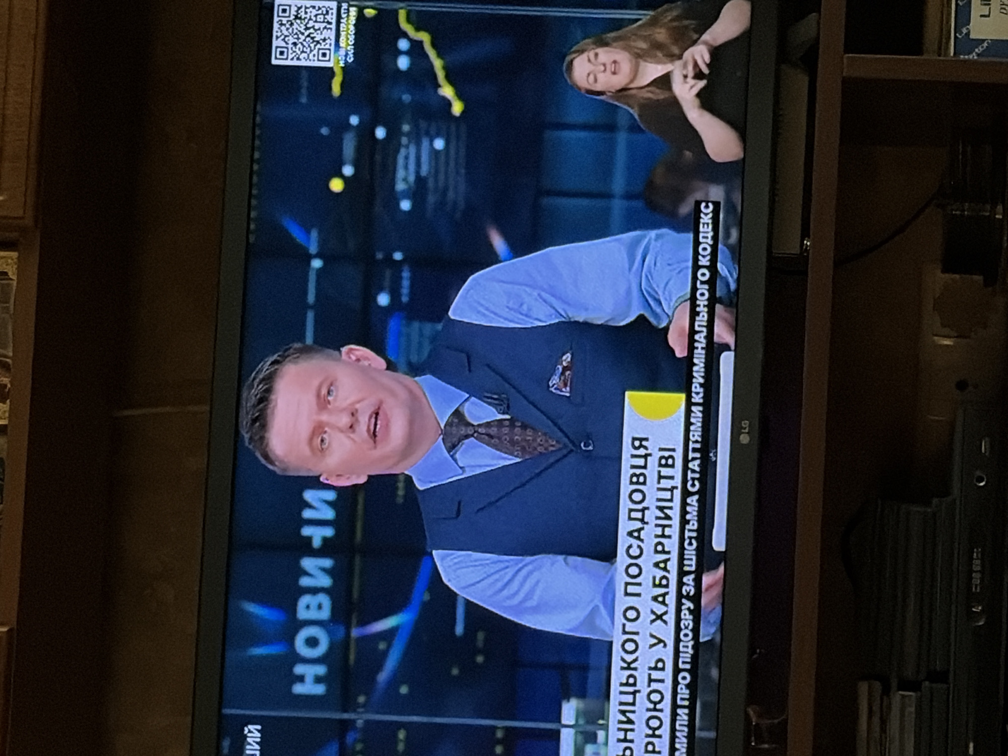 The image shows a television screen displaying a news broadcast. A male news anchor is speaking on camera, wearing a blue shirt and a dark vest. Text overlays on the screen are in Ukrainian and appear to relate to a news story. To the right of the anchor, a female individual is visible, possibly a guest or co-host, with her hands gesturing. The background of the broadcast features a city skyline at night, with glowing lights and abstract graphics. A QR code is also visible in the top right corner of the screen. The television is placed on a stand, with shelves below containing what appear to be media storage devices. The overall environment suggests an indoor setting, likely a living room or den.
