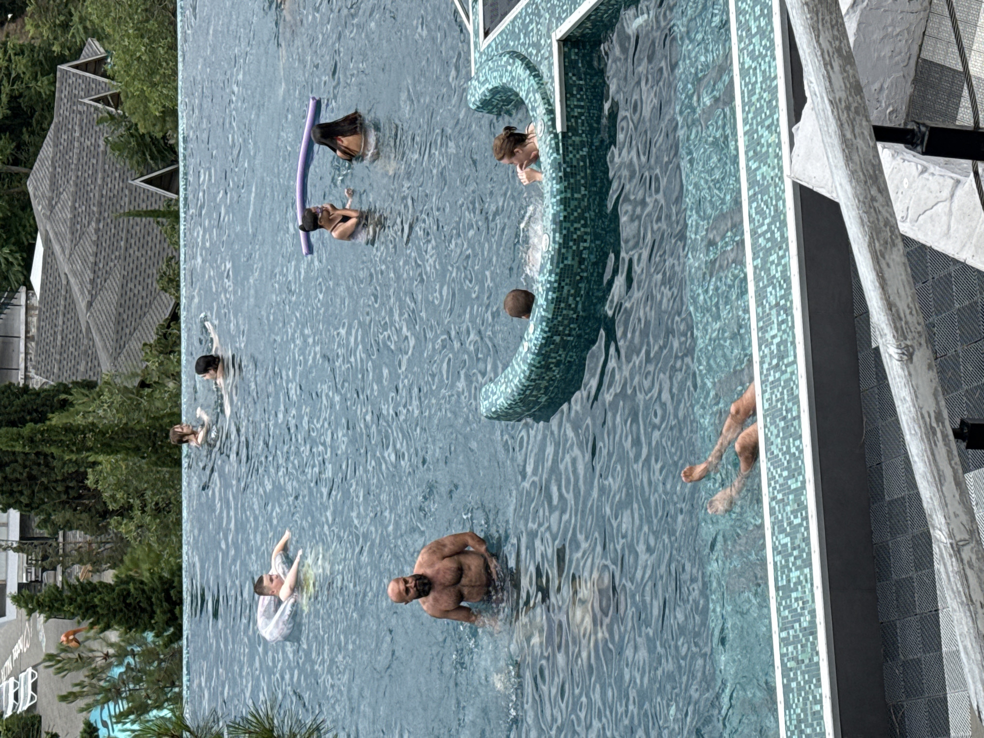 A wide shot captures an infinity pool with several people engaged in various aquatic activities. In the foreground, two pairs of legs are visible above the water, suggesting people are submerged within the pool. To the left, a man with a beard and muscular build stands in the water. Further back and to the left, a young person floats on a ring. In the middle distance, two young women are seen in the water, one holding a purple pool noodle. To the right and slightly further back, a woman is partially submerged in the pool. The pool itself is surrounded by a tiled edge and overlooks a forested hillside with buildings and rooftops in the background, characteristic of the cityscape of Yalta, Ukraine. The overall impression is of a resort or private residence with recreational amenities.