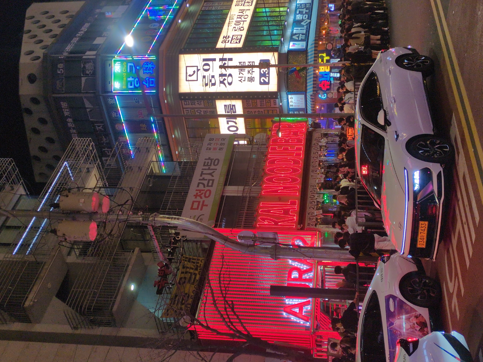 The image captures a nighttime scene in Seoul, South Korea, densely packed with neon lights and signage characteristic of the city's vibrant nightlife districts. The buildings are multi-storied, adorned with illuminated Korean characters and English text, advertising various businesses, including a "KAL NOODLE BAR" and a "Billiard Club". A notable red LED sign for "AR" is prominently displayed. The street is lined with people, many gathered on the sidewalks, suggesting a lively atmosphere. Parked vehicles are visible along the roadside, including a white car with Korean license plates. Power lines and utility poles are also present, adding to the urban environment.