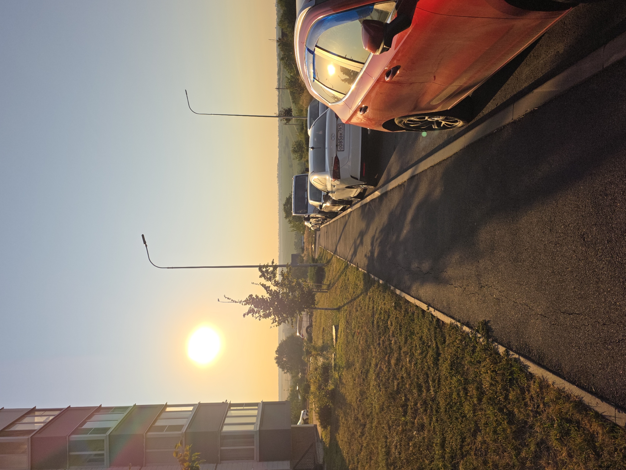 The image captures a street scene at sunrise in Rostov-on-Don, Russia. A multi-story building with a light-colored facade and windows dominates the left side of the frame. A paved walkway runs alongside the building, bordered by a grassy area. Several cars are parked along the walkway, including a red vehicle in the foreground that reflects the rising sun. A streetlamp stands on the right side of the walkway. The background features a treeline and rolling hills under a clear sky with a soft orange and yellow hue from the sunrise. The overall atmosphere is tranquil, depicting the early morning in a residential urban area.