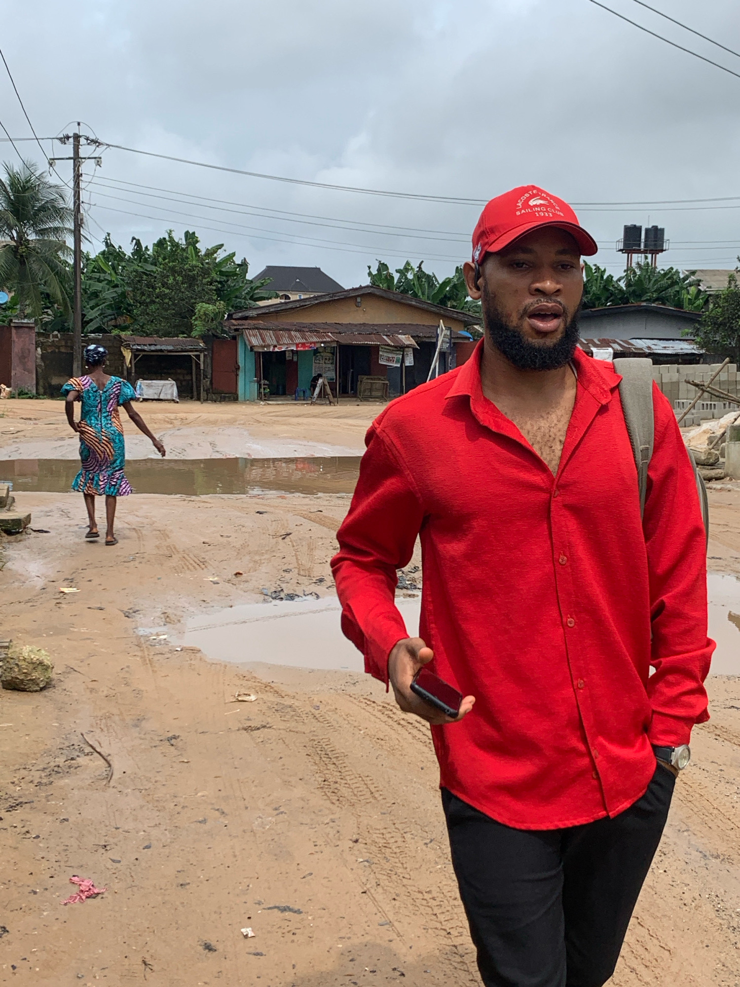 A man wearing a red collared shirt, black pants, and a red baseball cap stands in the foreground, facing slightly to the left. He holds a black smartphone in his left hand. Behind him, a woman in a colorful patterned dress walks away from the camera. The scene appears to be a muddy street in Umuode, Nigeria, with puddles of water on the ground. Several buildings, some appearing to be shops, are visible in the background. Overhead, electrical wires stretch across the frame, supported by a utility pole. The sky is overcast.