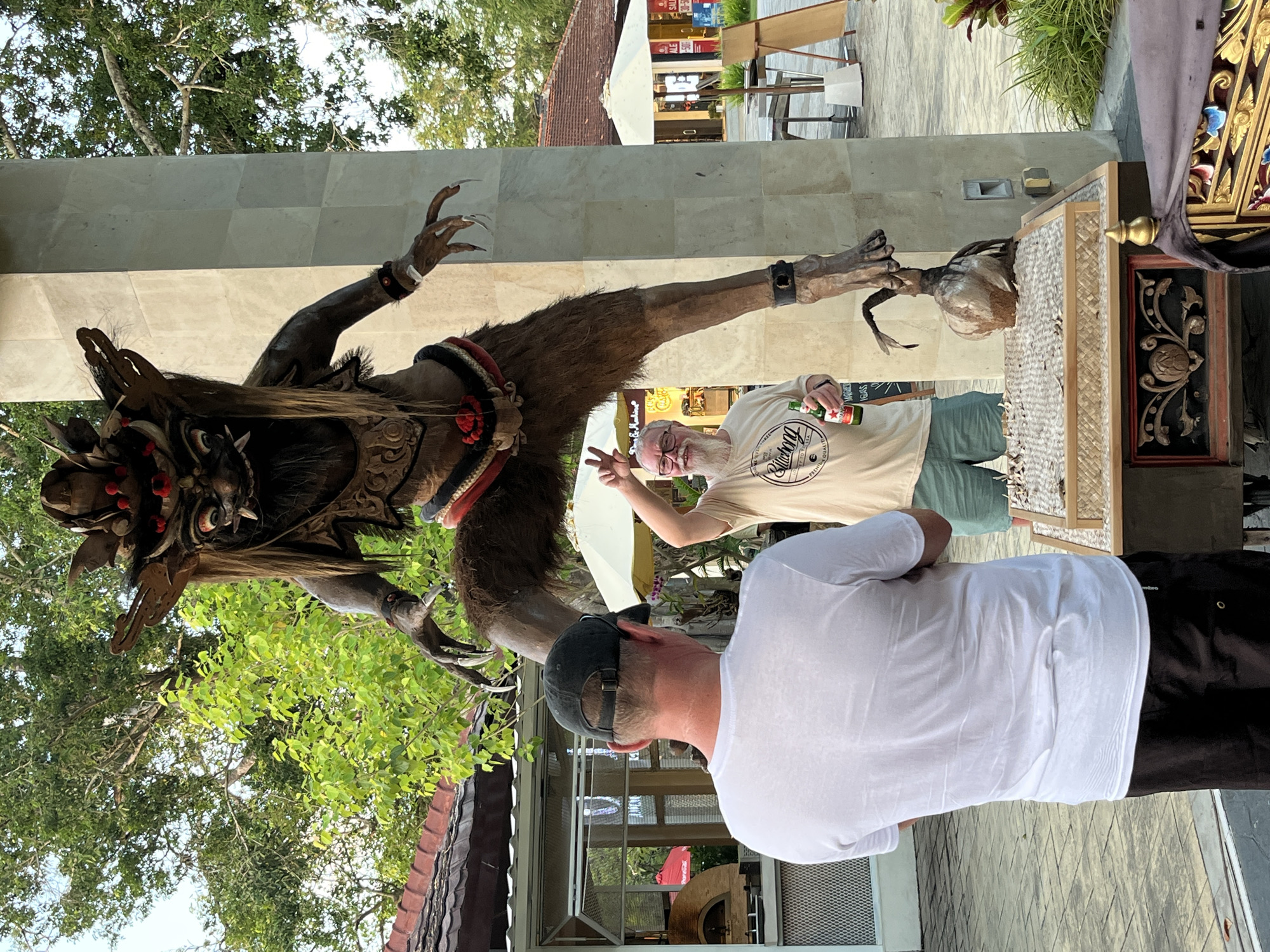 In Kuta Selatan, Indonesia, two men stand near a large, imposing Balinese demon statue. The statue, dark brown and intricately carved, features sharp claws, long hair, and a fierce expression. It is positioned with its legs spread wide, straddling a stone pillar. One man, wearing a white t-shirt and a dark baseball cap, stands with his back to the camera, facing the statue. The other man, with a white beard and glasses, wears a light-colored t-shirt and shorts. He holds a bottle of beer in one hand and makes a peace sign with the other. He is looking towards the statue. In front of the men and the statue's base, a small, ornate wooden altar is visible, covered with what appears to be pebbles. The background shows lush green trees and parts of buildings, suggesting an outdoor public space or entertainment area, possibly a market or street. The lighting indicates it is daytime.