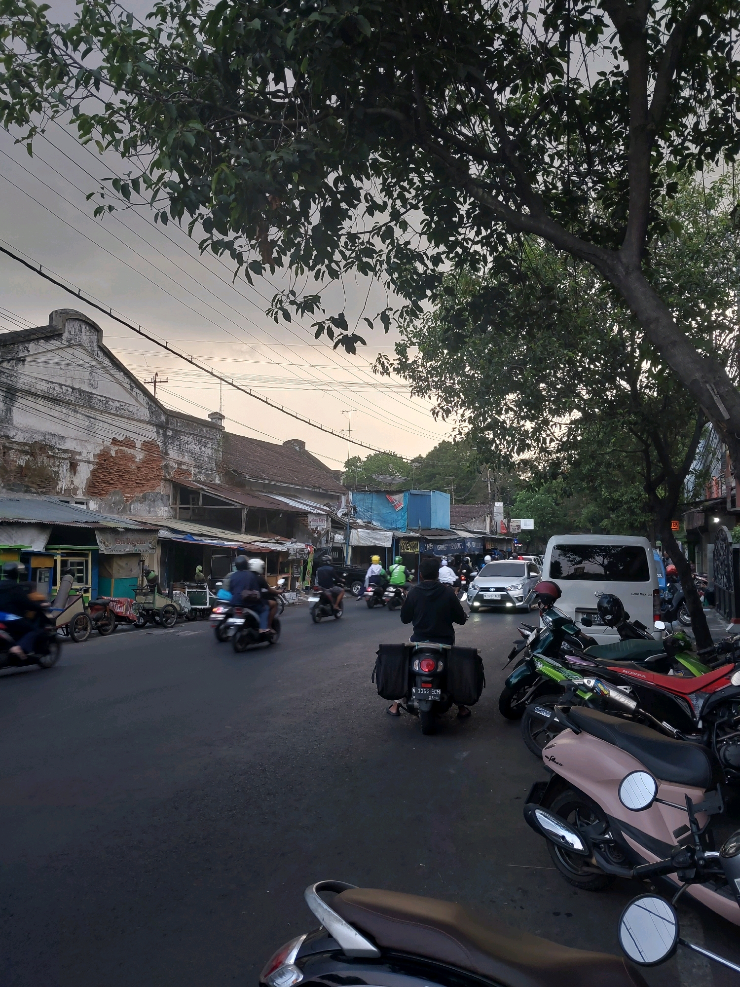 This image depicts a street scene in Malang, Indonesia, likely during the late afternoon or early evening, given the soft, overcast lighting. The street is paved and appears to be a busy thoroughfare with significant motorcycle traffic. On the left side of the street are low-rise buildings with aged facades, some showing exposed brickwork, suggesting an older part of the city. These buildings house various small businesses, indicated by signage and awnings.
Numerous motorcycles are present, both in motion and parked along the roadside. Several individuals are riding motorcycles, with some wearing helmets. In the foreground, several motorcycles are parked. A white van is also visible on the right side of the street. The environment is characterized by overhead power lines and trees lining the street, casting shadows. The overall impression is of a bustling urban environment in Indonesia.