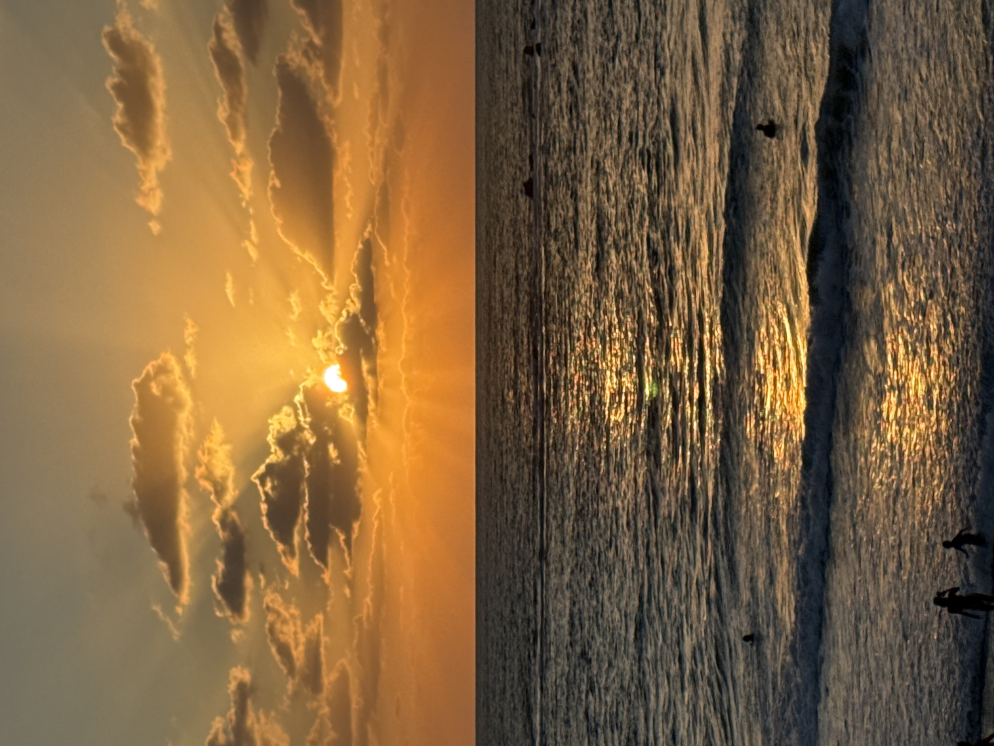 A vertical, close-up, high-angle shot captures a vibrant sunset over the Mediterranean Sea in Tel Aviv, Israel. The sky is a rich tapestry of orange and yellow hues, with scattered clouds illuminated by the setting sun. Rays of light pierce through the clouds, creating a dramatic effect. The sun itself is partially obscured by clouds, casting a golden glow on the water.
The sea is dark blue, reflecting the warm colors of the sunset. Gentle waves break near the shore, and the water sparkles with sunlight. Several small figures are visible in the water, including swimmers and possibly surfers. In the foreground, silhouettes of people are seen on the sandy beach, enjoying the evening. The overall scene depicts a peaceful and picturesque coastal sunset in Tel Aviv.