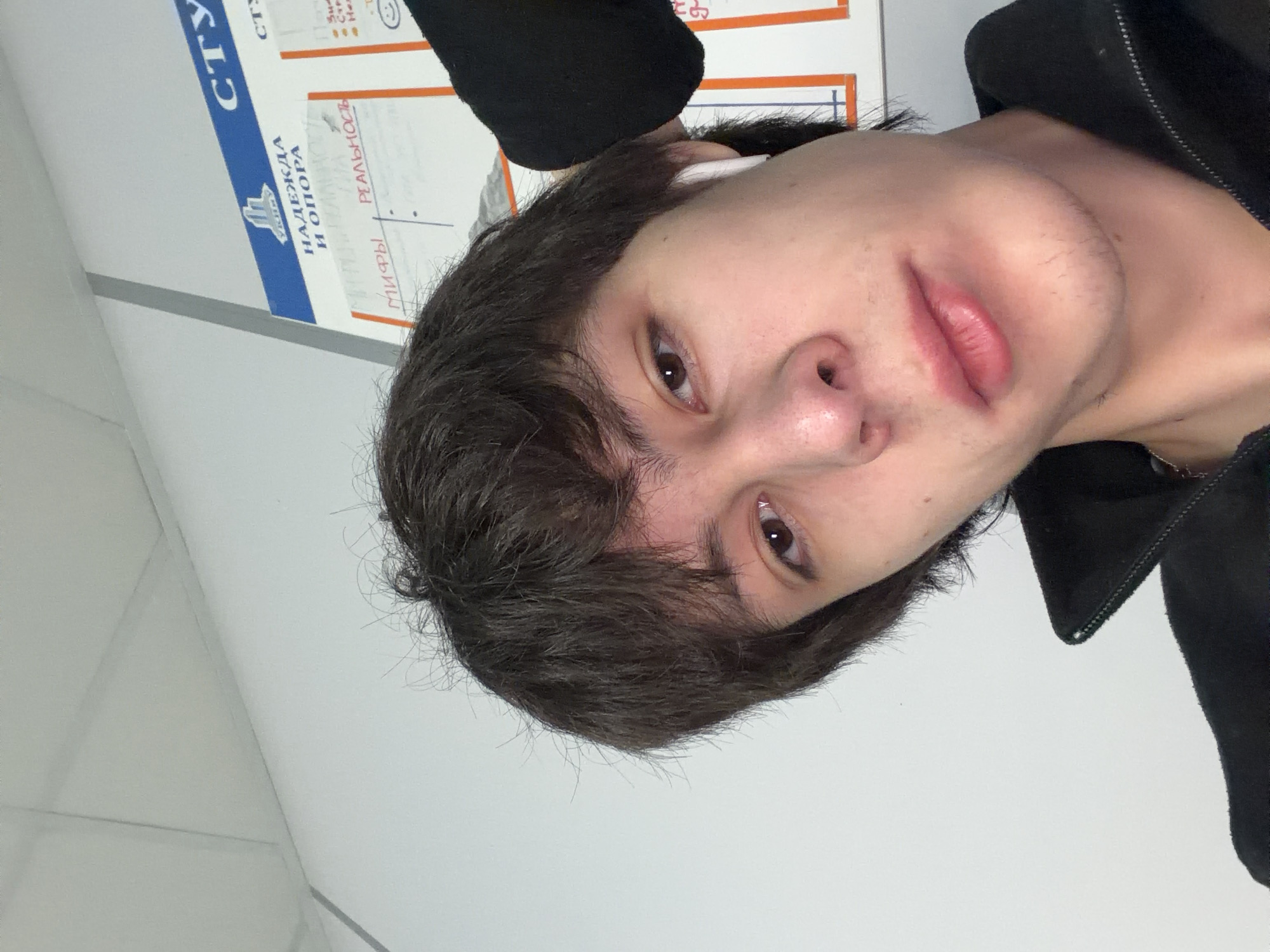 The image is a selfie taken by a young male with dark, messy hair and brown eyes. He is wearing a black jacket and white wireless earbuds. In the background, a white wall is visible with a poster that has Russian text and diagrams. One visible section of the poster reads "НАДЕЖДА И ОПОРА" (Hope and Support) and another section has the word "СТУ" partially visible. There are also handwritten notes on the poster. The lighting suggests an indoor environment. It is not possible to determine the specific location in Yekaterinburg from the image alone.