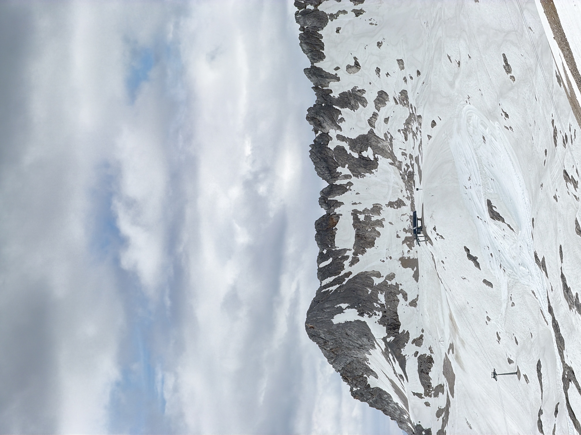 This is a vertical, medium-shot photograph of the Zugspitze mountain in Garmisch-Partenkirchen, Germany, on a cloudy day. The mountain is covered in snow and patches of rock. In the foreground, a ski lift is visible, with its cables stretching across the snowy slope. The ski lift station, a modern structure with a blue roof, is situated further up the mountain. The sky is filled with a mix of gray and white clouds, with glimpses of blue sky peeking through. The overall scene depicts a vast, rugged mountain landscape, typical of a winter resort or a high-altitude alpine environment.