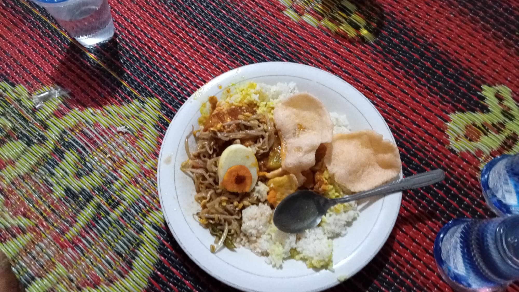 A white plate filled with food sits on a red, black, and yellow woven mat. The plate contains white rice, bean sprouts with sauce, a hard-boiled egg cut in half with the yolk visible, and two pieces of puffed crackers. A spoon rests on the plate, partially submerged in the rice. To the left of the plate, a partially visible clear plastic cup filled with water is on the mat. To the right of the plate, two blue and white plastic bottles are also on the mat. The scene appears to be an outdoor setting, possibly a meal in Uteunghatom, Indonesia.