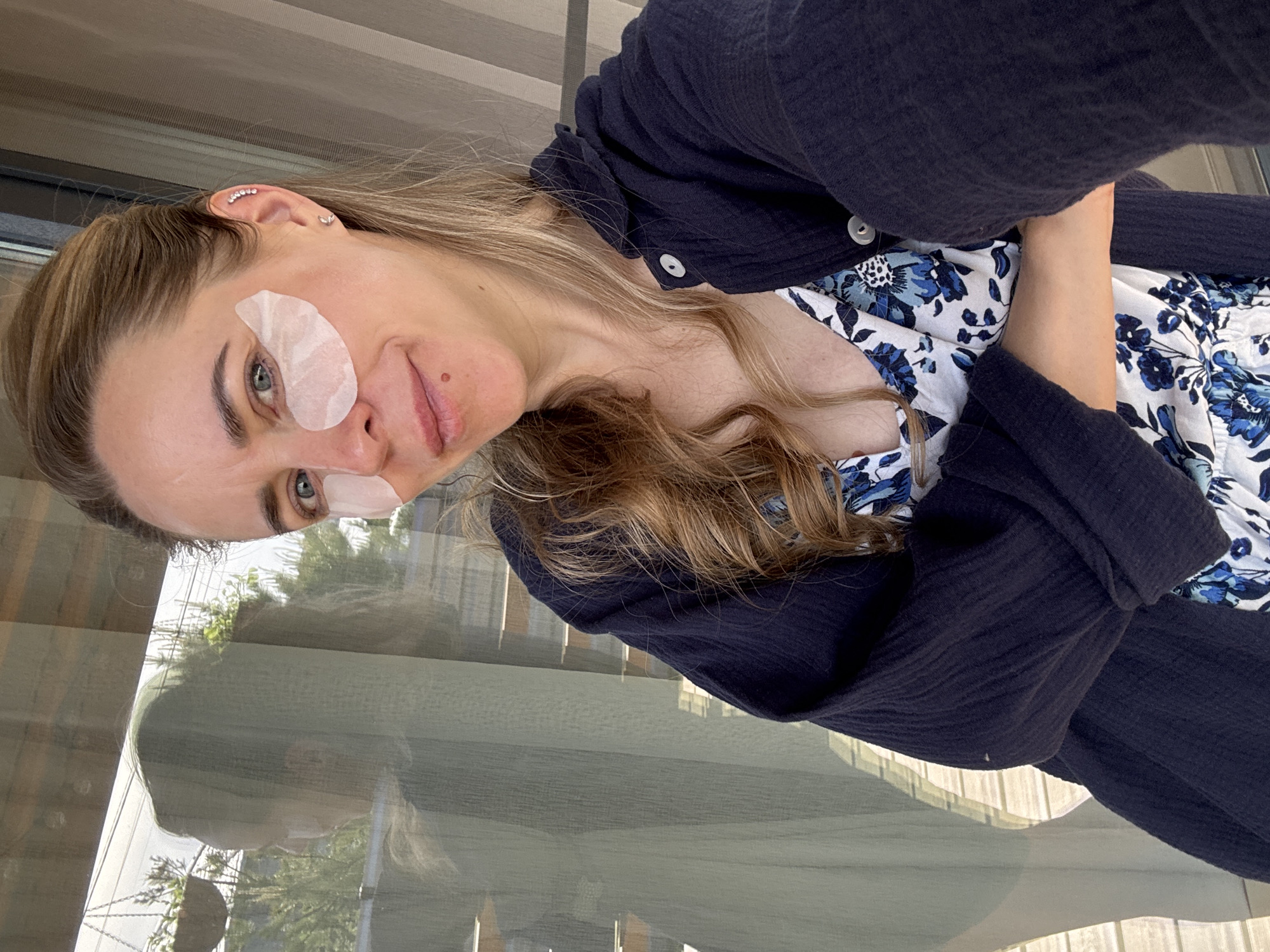 A fair-skinned woman with light brown hair is looking into the camera at a downward angle. She has under-eye patches on both eyes. She is wearing a floral-print dress with a dark navy blue overshirt. Behind her is a glass panel that reflects vegetation. The location is suggested to be in Chaikovsky, Russia, but this cannot be definitively confirmed from the image alone.