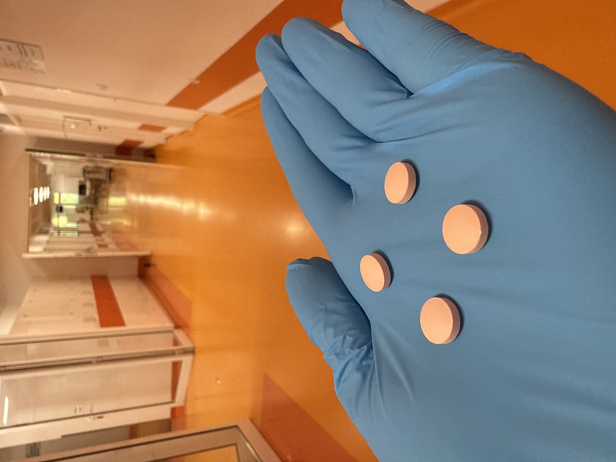 A close-up shot shows a gloved hand, wearing a blue nitrile glove, holding four round, light pink pills on the palm. The background is a brightly lit hospital corridor with an orange floor and white walls. The corridor recedes into the distance, with doorways visible on either side and fluorescent lighting overhead. The image appears to have been taken in a hospital setting, likely in Pleven, Bulgaria, given the context of the prompt.