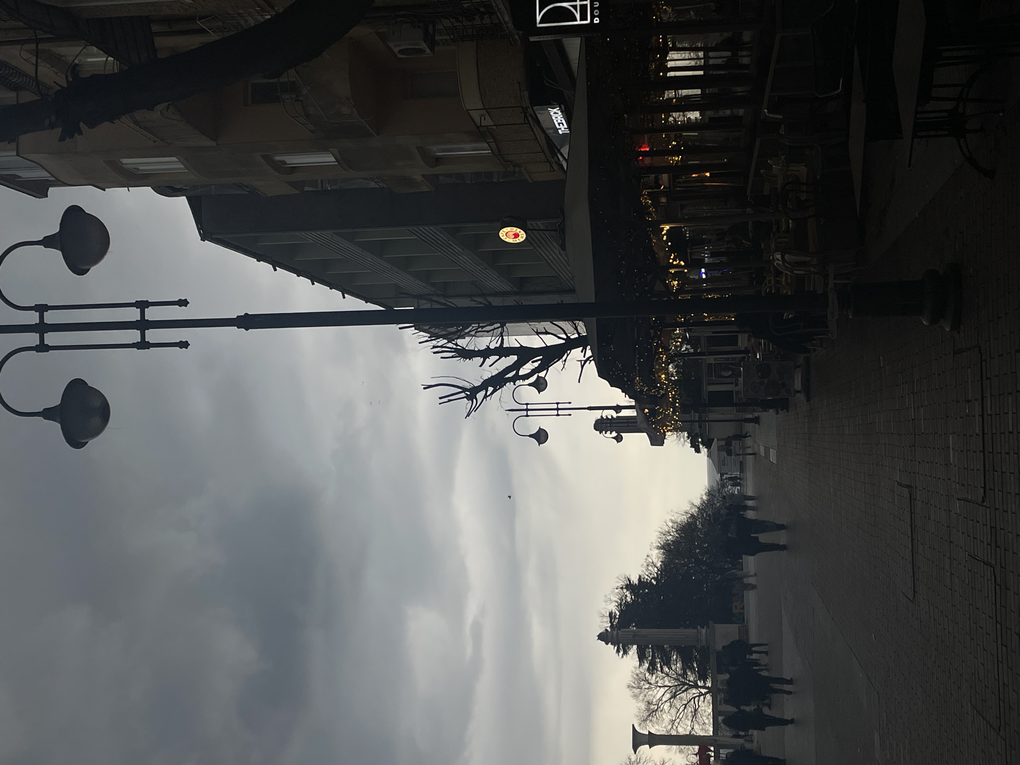 The image shows a street scene in Varna, Bulgaria on a cloudy day. A row of buildings lines the street, with visible signage for businesses like "THE BRICK" and "DOUBLE." Lamp posts with distinctive, rounded lamps are placed along the sidewalk. People are walking along the paved pathway. Outdoor seating and tables are visible along the building facades. Trees line the opposite side of the sidewalk. The overall light is low due to the overcast sky.