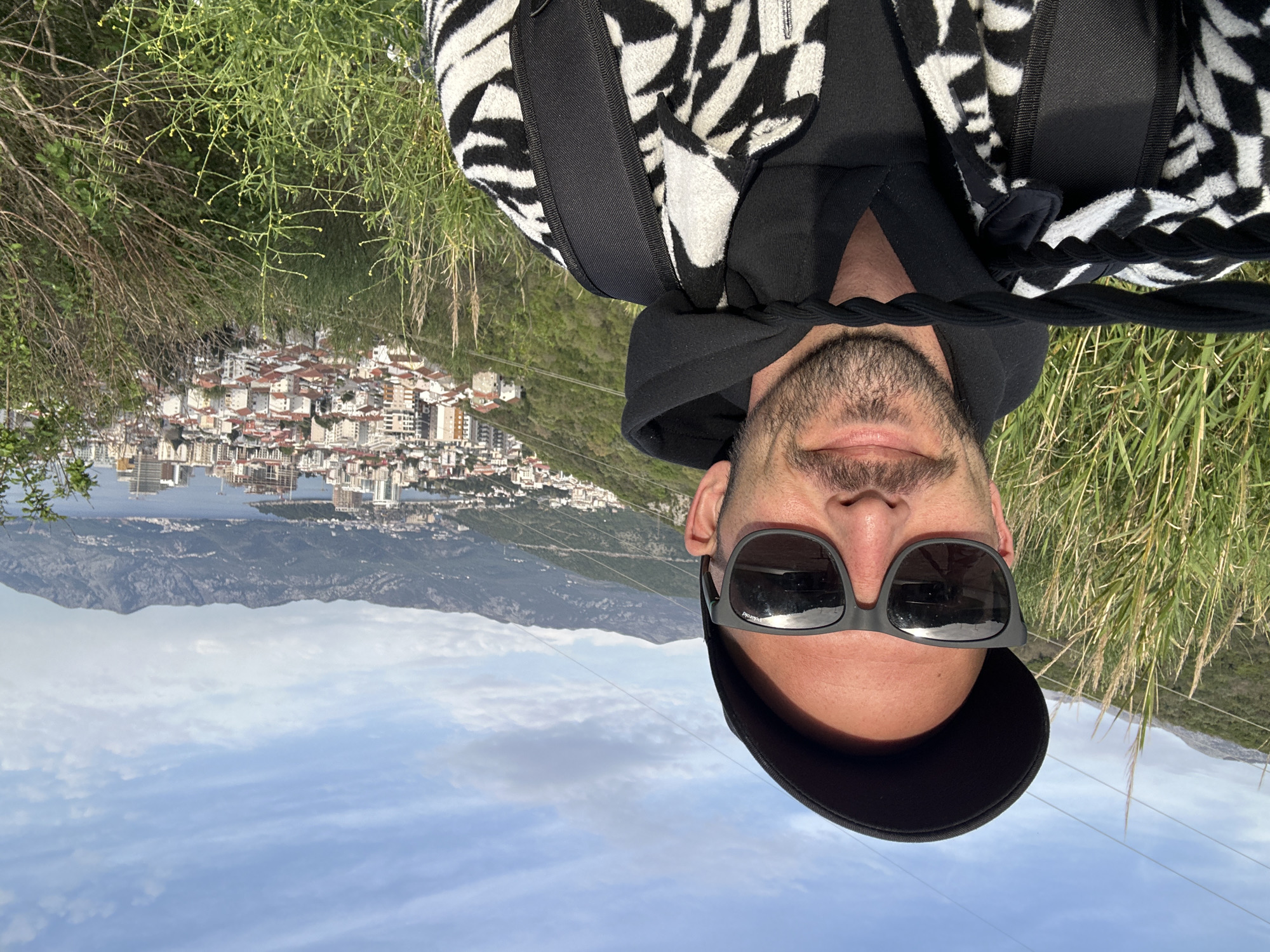 A man is taking a selfie. He is wearing sunglasses and a black cap. He has a beard. He is wearing a black and white patterned jacket. He is standing on a hill overlooking the city of Budva, Montenegro. The sky is blue with some clouds. There are mountains in the background. There is a body of water behind the city. There is some green vegetation.