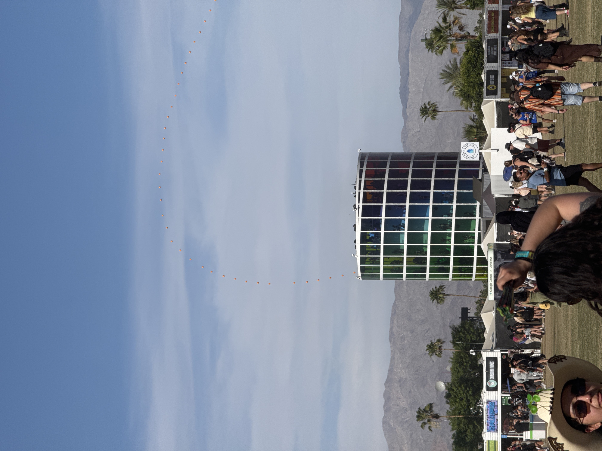 Here is a detailed description of the image:
**Location:** Indio, California, United States of America (likely at the Coachella Valley Music and Arts Festival).
**Scene Description:** The image presents an outdoor event, possibly a music festival, under a clear sky. An art installation constructed of translucent rainbow-colored panels is prominent in the center of the frame, surrounded by a large crowd of people. The crowd is congregated on a grassy field, with multiple vendor booths. Palm trees dot the landscape behind the crowds, and a mountain range is visible in the background. The presence of people wearing festival attire, vendor booths selling food, merchandise, and the presence of an art installation confirm the festival setting. Above, a string of what appears to be orange, spherical objects spans the sky in a curved line. A woman with a straw hat is also in the frame.