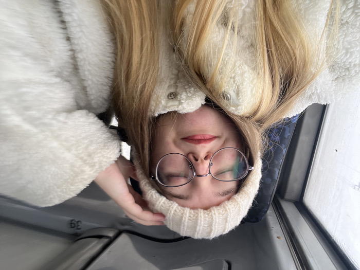 A young adult female, with long blonde hair, fair skin, and round wire-rimmed eyeglasses, is depicted inside what appears to be a public transport vehicle in the city of Липицы, Russia. She is wearing a cream-colored knit beanie and a white, textured, possibly sherpa-style jacket. Her eyes are closed, and her lips are slightly parted in a subtle smile, adorned with light pink lipstick. Her right hand is partially visible, touching the top of her beanie on the right side of her head. The background consists of a dark blue patterned seat and a window to her left, which is obscured by condensation or frost, showing an indistinct bright, possibly wintery, outdoor environment. Above her head, a section of the vehicle's gray interior ceiling or overhead compartment is visible, bearing the alphanumeric marking "3g".