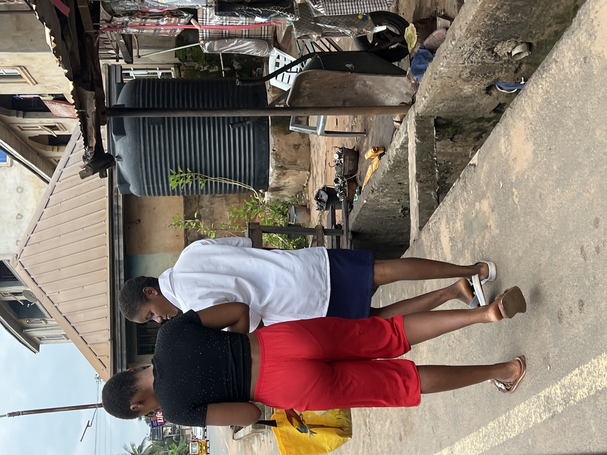 Two young women are walking on a sidewalk in Nkpor, Nigeria. The woman in the foreground on the left is wearing a black t-shirt and red shorts. She is carrying a yellow bag in her left hand. The woman on the right is wearing a white shirt and a dark blue skirt. They are walking away from the camera. The sidewalk is made of concrete. Behind them, there is a building with a brown roof. To the right of the building is a large black water tank. Further to the right, there are stalls with goods displayed. The environment appears to be a busy urban area.