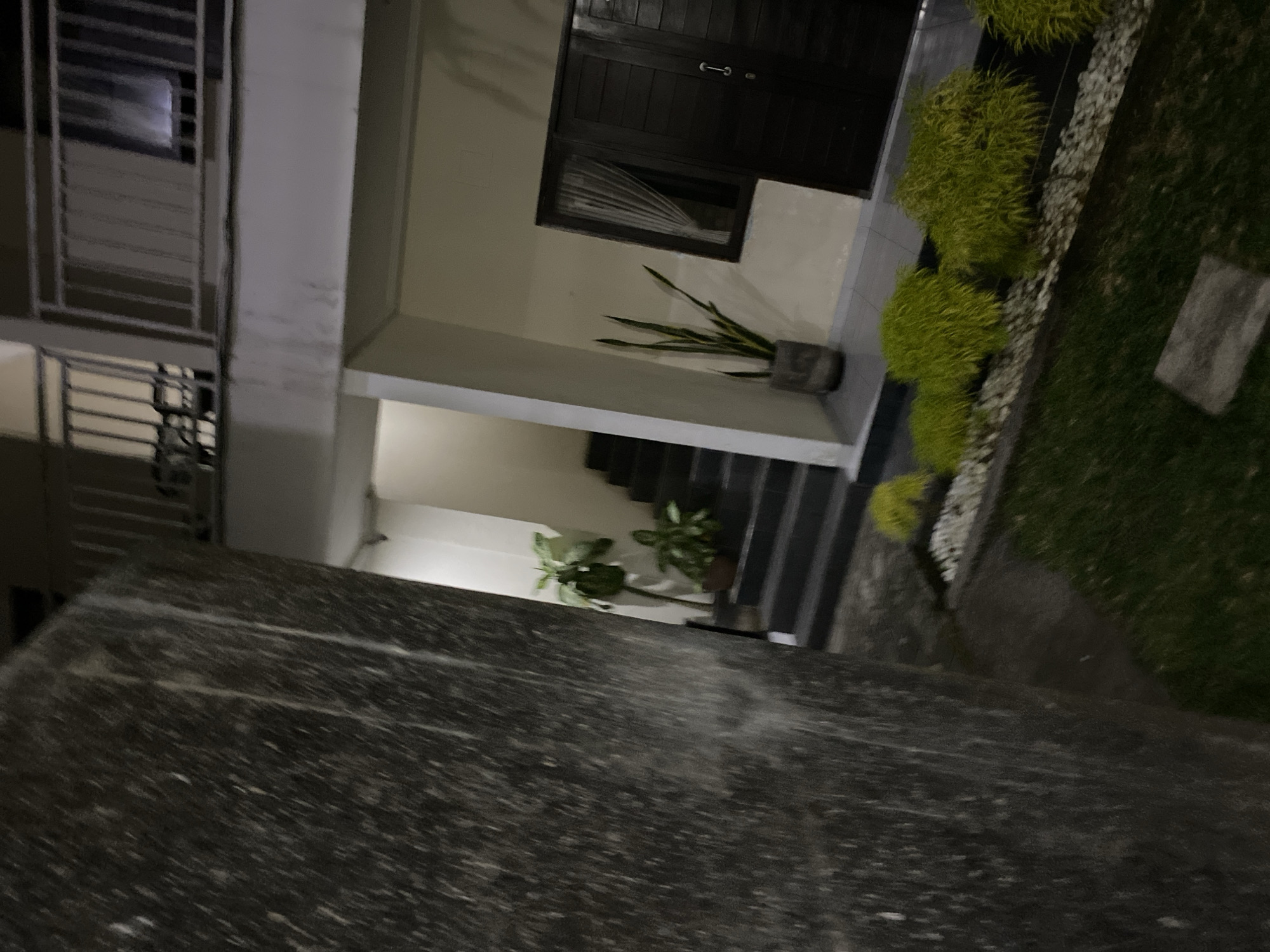 This image depicts an outdoor area at night, possibly a residential or commercial building. In the foreground, a large, textured dark gray wall is visible on the left, partially obscuring the scene. To the right of the wall, a set of dark gray stairs leads upwards. Several potted plants are placed along the stairs and near the entrance of a building. The building itself has a light-colored facade and a dark brown double door with a silver handle. A window with sheer white curtains is situated next to the door. In the lower right portion of the image, a garden bed with bright green, bushy plants and white decorative stones is visible, bordered by a patch of green grass. Above the building, a balcony with a white railing can be seen. The lighting suggests it is nighttime, with artificial lights illuminating the area. The environment appears to be a well-maintained outdoor space, possibly part of a resort or hotel in Kuta Selatan, Indonesia, given the tropical vegetation and architectural style. There are no people visible in the image.
